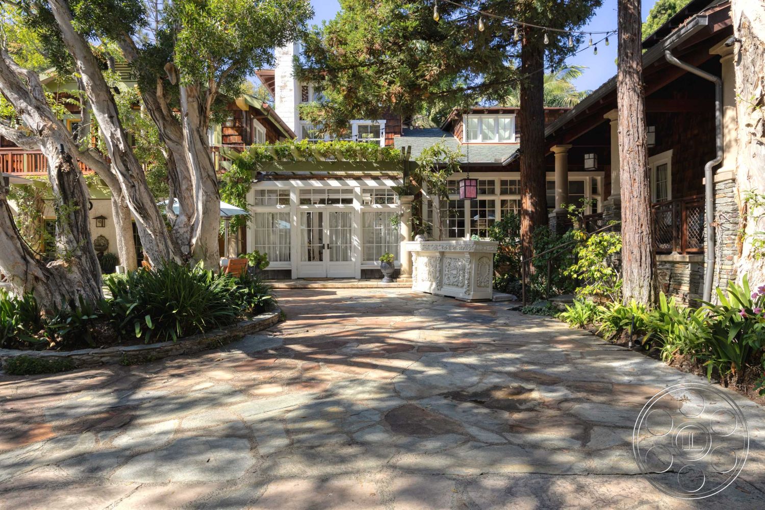 Americana 48 - exterior, outdoor seating area, exposed wood beams, gabled roof, evergreen trees, stone patio flooring, lush garden landscaping, tranquil garden setting, white wooden window frames, decorative stone fountain, flagstone patio area, wooden garden trellis, traditional cottage-style home, dark wood shingles