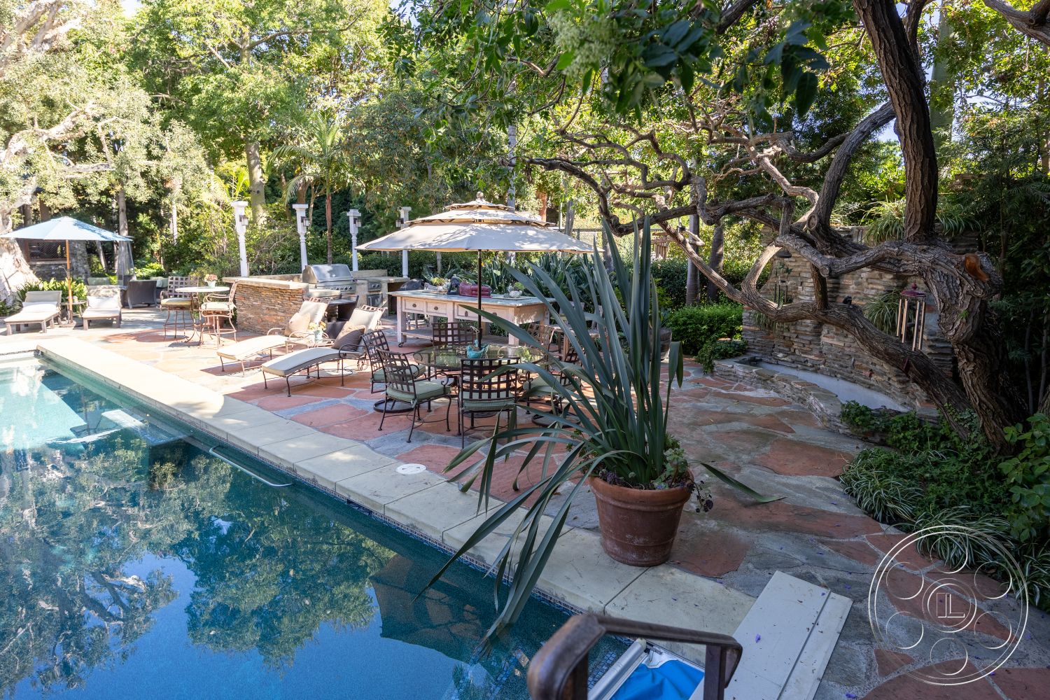 Americana 48 - exterior, stone accent wall, potted plants, natural wood beams, rectangular pool, outdoor kitchen area, stone patio flooring, concrete pool border, lush green surroundings, flagstone pool deck, high back patio chairs, covered patio with umbrella, built-in grill station, multi-tiered landscaping, freestanding outdoor seating, shaded lounging area