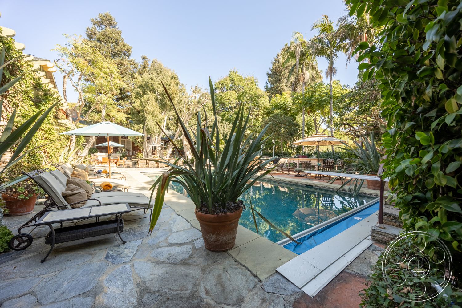 Americana 48 - exterior, potted plants, outdoor seating area, garden patio, tropical setting, outdoor lounge chairs, lap pool, rectangular pool, wooden deck area, stone patio flooring, umbrella shade structures, lush tree landscaping, aluminum pool railings, circular pavers