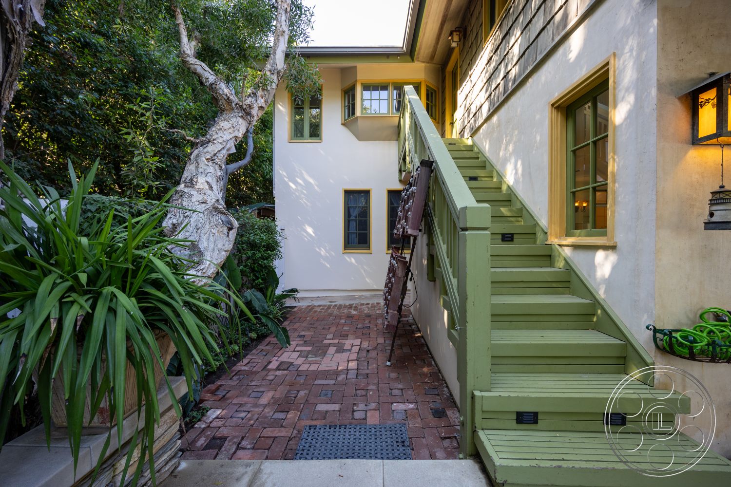 Americana 48 - exterior, concrete wall, brick walkway, wooden stairs, overhanging eaves, textured plaster wall, wood shingle siding, green painted wooden steps, yellow painted window frames, leafy landscaping