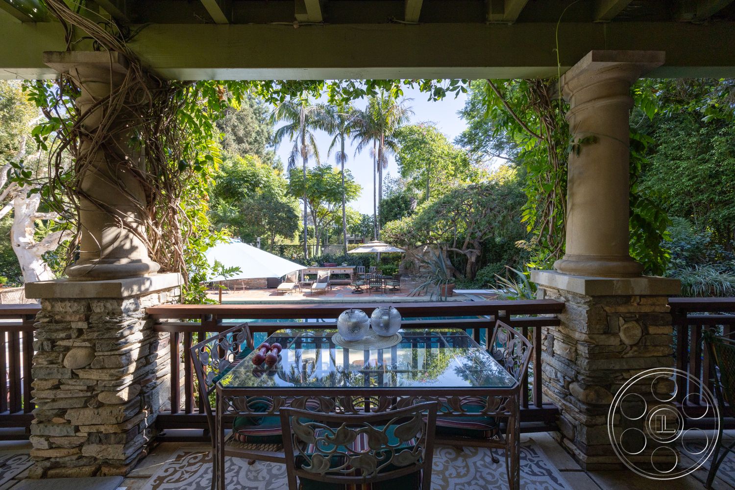 Americana 48 - exterior, palm trees, outdoor seating area, glass dining table, lush tropical landscaping, wooden railing, stone column bases, decorative stone veneer, textured tile patio flooring, sunny poolside view, covered outdoor area, sloped garden, vining plants, pool deck area