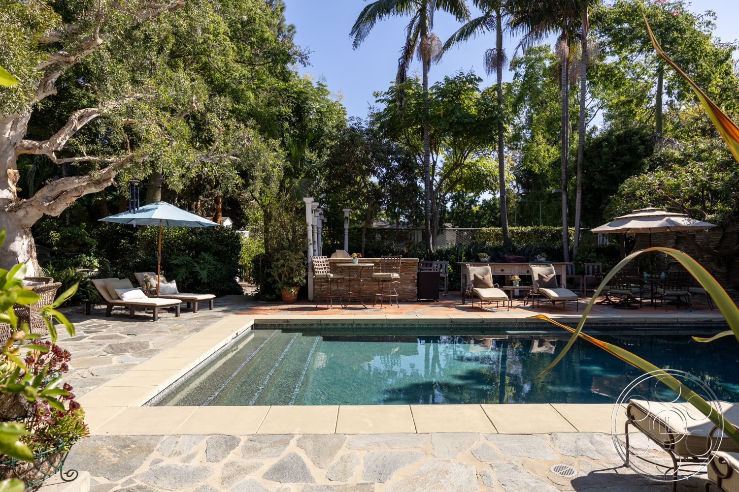 Americana 48 - exterior, surrounding greenery, rectangular swimming pool, outdoor lounge area, lush tropical landscaping, tall palm trees, stone patio flooring, concrete pool coping, natural stone pool deck, wooden sun loungers, umbrella over seating area, brick barbecue grill, tile waterline around pool