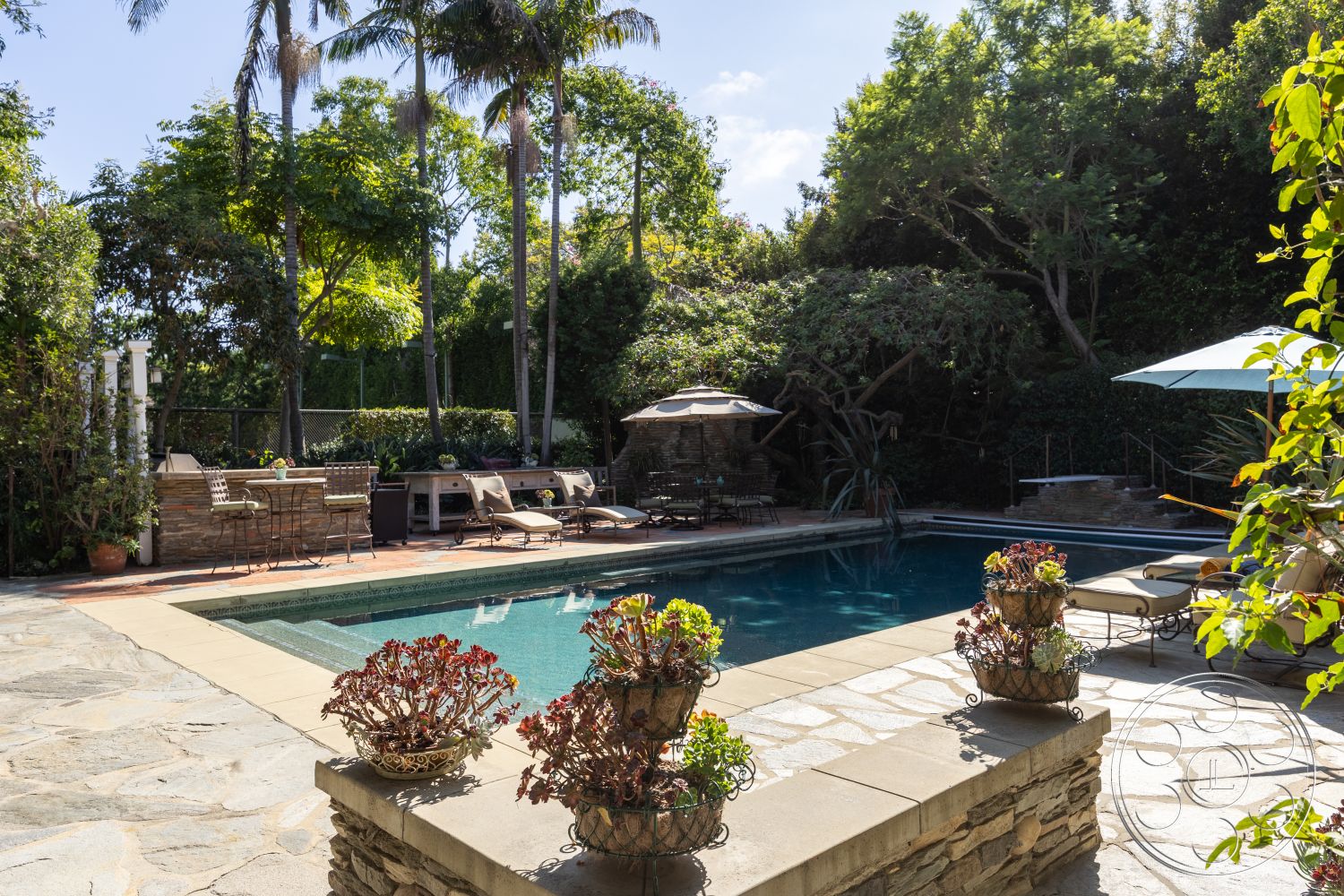 Americana 48 - exterior, covered patio, rectangular pool, stone pool deck, landscaped garden area, fenced backyard, concrete pool border, natural stone patio flooring, lush greenery surrounding the pool, built-in outdoor kitchen, outdoor living area with chairs, potted plants with succulents, sun loungers with cushions, umbrella for shade