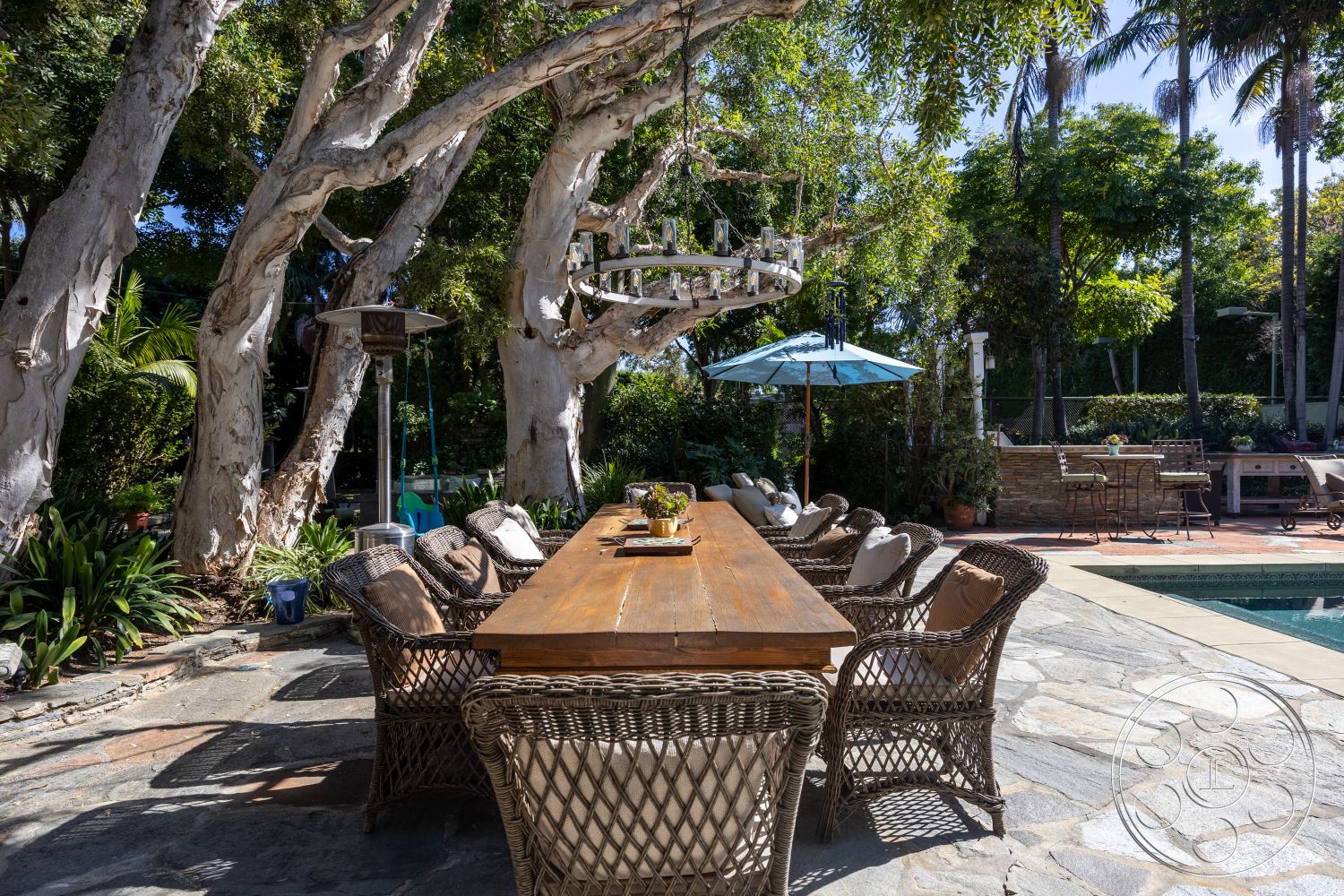 Americana 48 - exterior, tropical landscaping, in-ground swimming pool, outdoor umbrella, natural stone patio flooring, private backyard oasis, hanging chandelier lighting, sunny garden setting, rectangular wooden dining table, woven wicker dining chairs, multi-trunk eucalyptus trees, paver stone pool border, built-in outdoor kitchen area