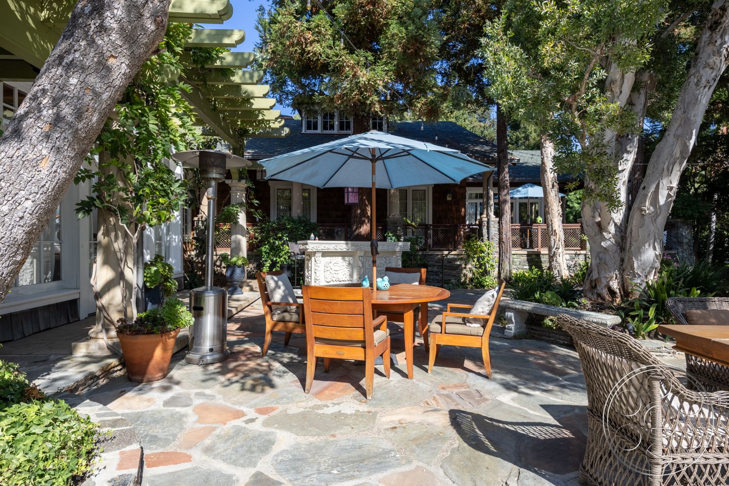 Americana 48 - exterior, wooden dining table, sunny outdoor setting, metal patio heater, landscaped garden area, lush greenery surrounding patio, natural stone patio flooring, wicker outdoor chairs, tile outdoor dining area, slate outdoor features, blue umbrella, attached pergola structure, brick accent wall of house, wooden beams of pergola