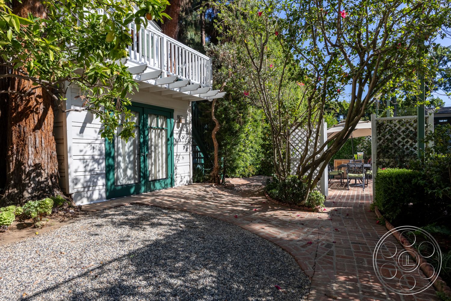 Americana 48 - exterior, gravel pathway, landscaped garden setting, brick patio flooring, sunny outdoor environment, wooden pergola structure, curved pathway design, wooden balcony railing, green double doors, white vertical siding walls, lush garden planting, flowering tree foliage, scenic outdoor dining area