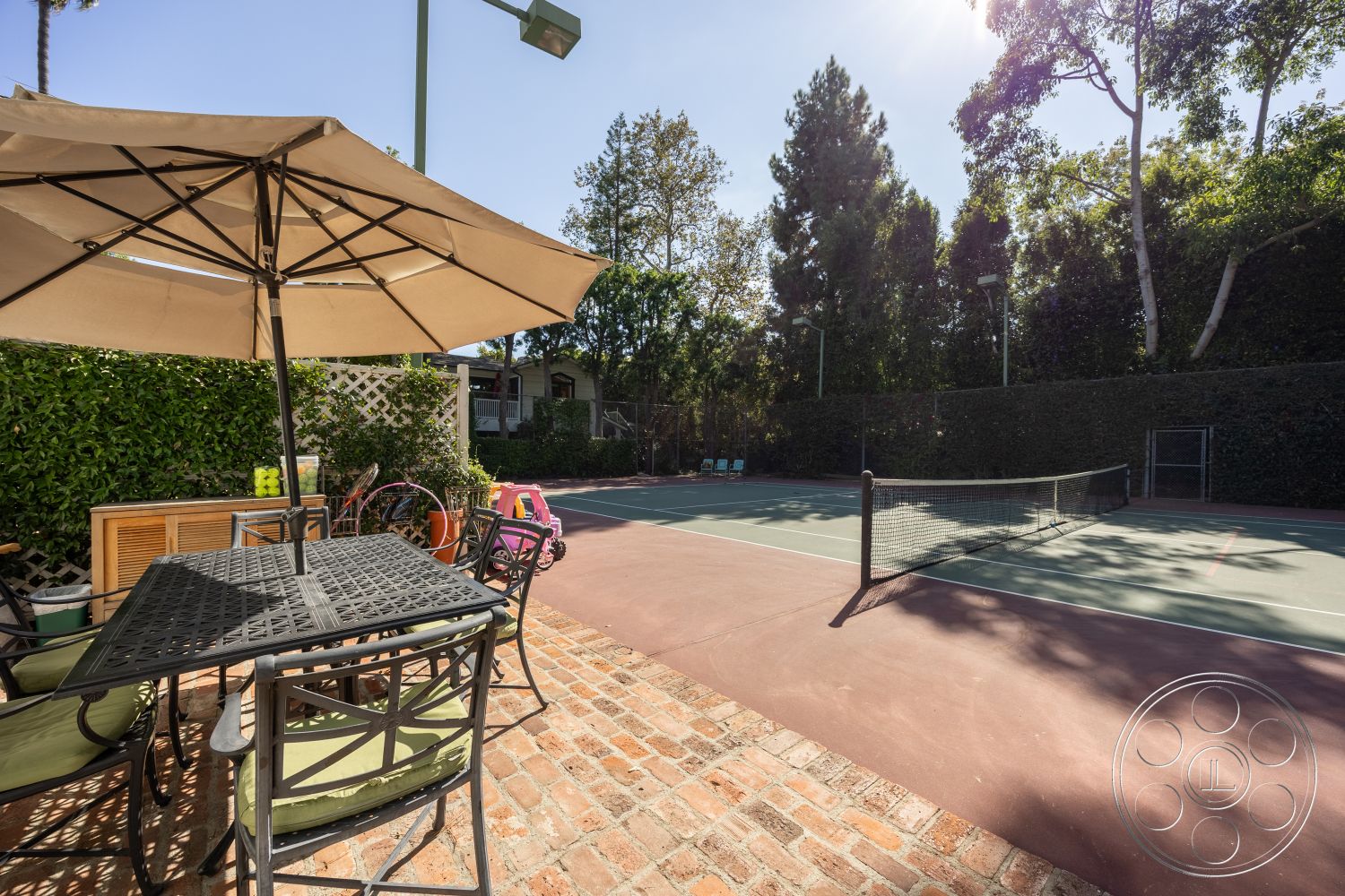 Americana 48 - exterior, lush green landscaping, green upholstered chairs, brick patio flooring, sunlit outdoor setting, light pole fixtures, metal outdoor dining table, canvas umbrella shade, tennis court with green surface, horizontally aligned wooden fence