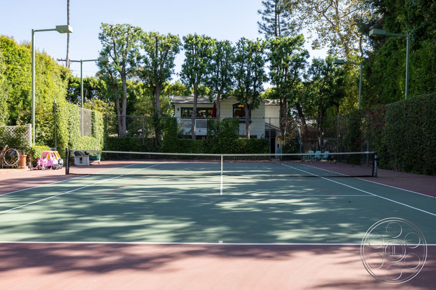 Americana 48 - residential backyard, surrounding hedge landscaping, asphalt tennis court surface, green tennis court lines, line of trees along back court, wooden exterior house visible, outdoor tennis court setting, daylight environment