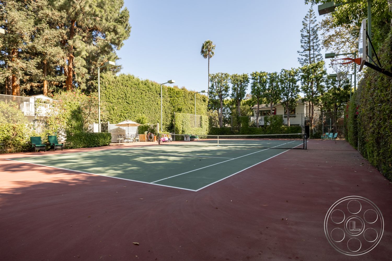 Americana 48 - exterior, outdoor seating, chain link fence, surrounding palm trees, wooden deck area, sunny backyard setting, landscaped hedge border, red clay tennis court surface, concrete tennis court base, outdoor basketball hoop