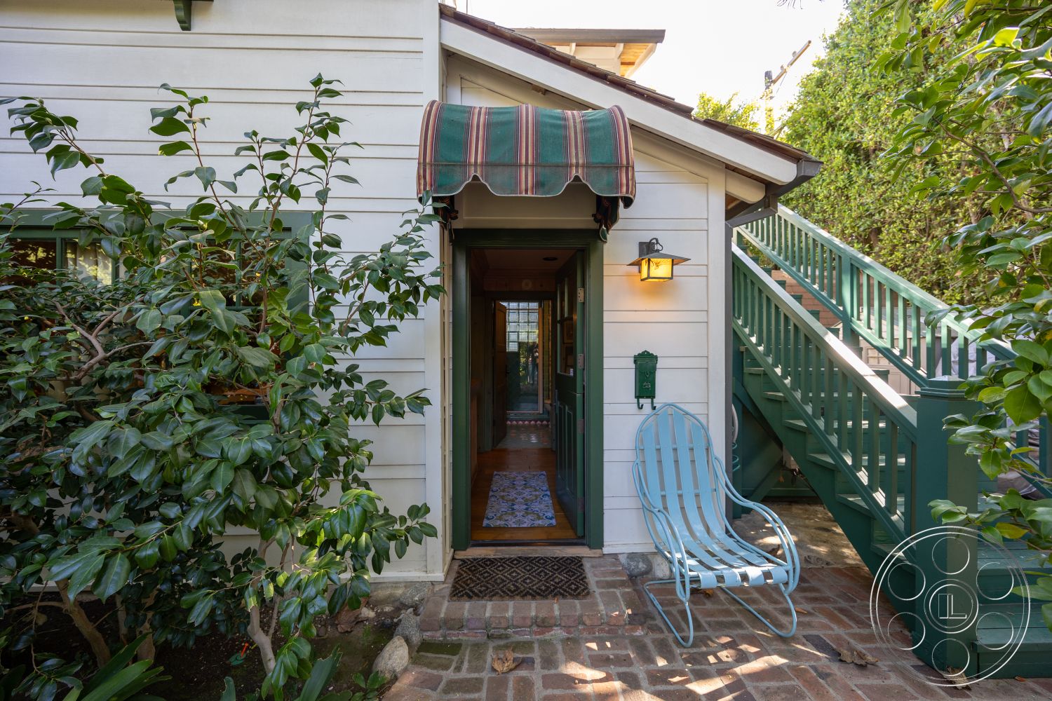 Americana 48 - exterior, landscaped garden, surrounding greenery, brick pathway, front porch entry, wood siding walls, striped canvas awning, green wooden stairs, blue metal chair