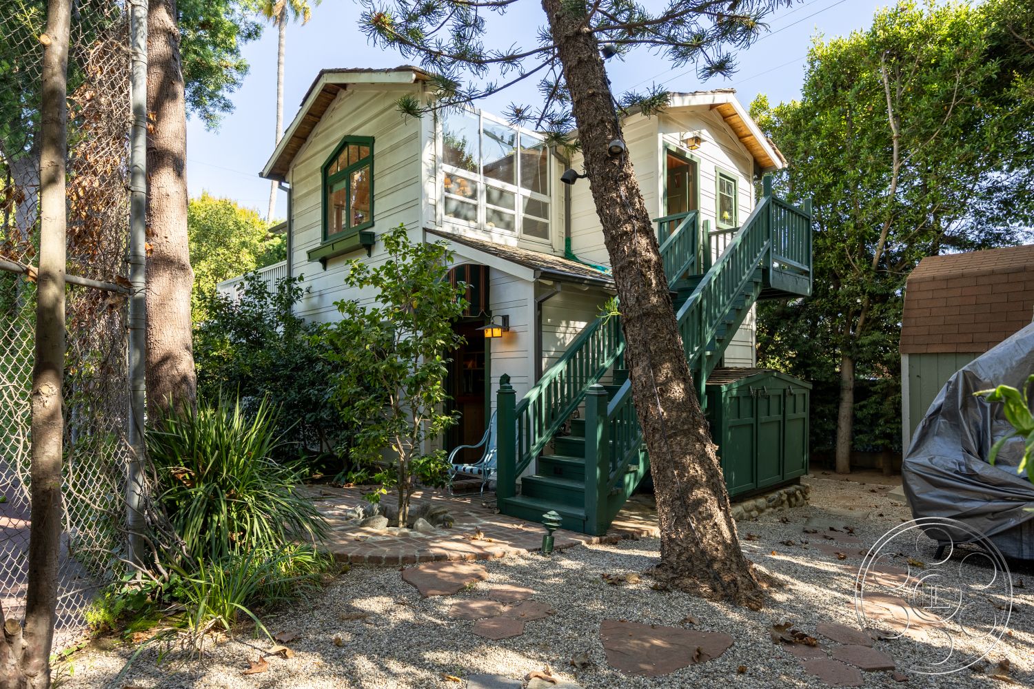 Americana 48 - exterior, outdoor seating area, wooden staircase, surrounding trees, stone pathway, gravel landscaping, planted shrubs, wood siding, double-hung windows, shingle roof, gable roof, green trim, wooden balcony railings