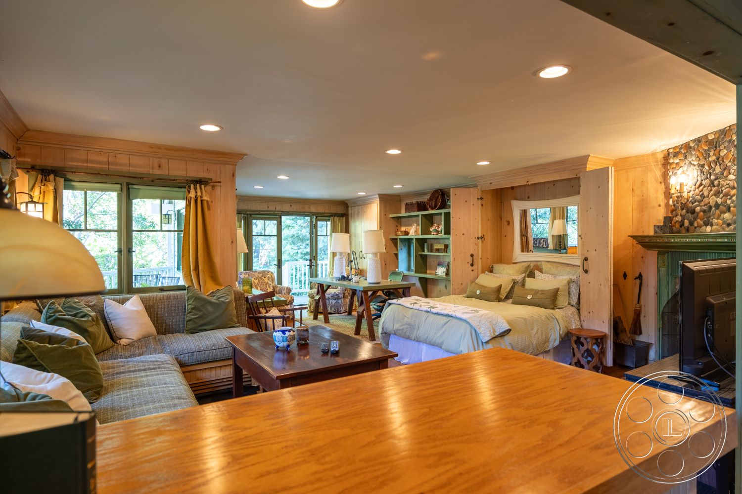 Americana 48 - large windows, natural light, exposed beam ceiling, open concept layout, wooden dining table, plush sectional sofa, hardwood floor, bedside table, textured throw blankets, green accent pillows, cozy reading nook, decorative shelves, stone fireplace surround, wood paneling walls