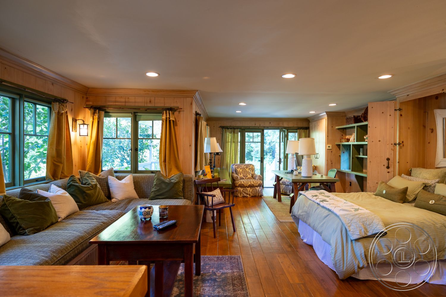 Americana 48 - large windows, natural light, hardwood flooring, wooden coffee table, open floor plan, cozy seating area, built-in shelving, vaulted ceiling, textured ceiling, soft bedding, plush throw pillows, comfortable armchair, green curtains, decorative lighting fixtures, wood paneling walls