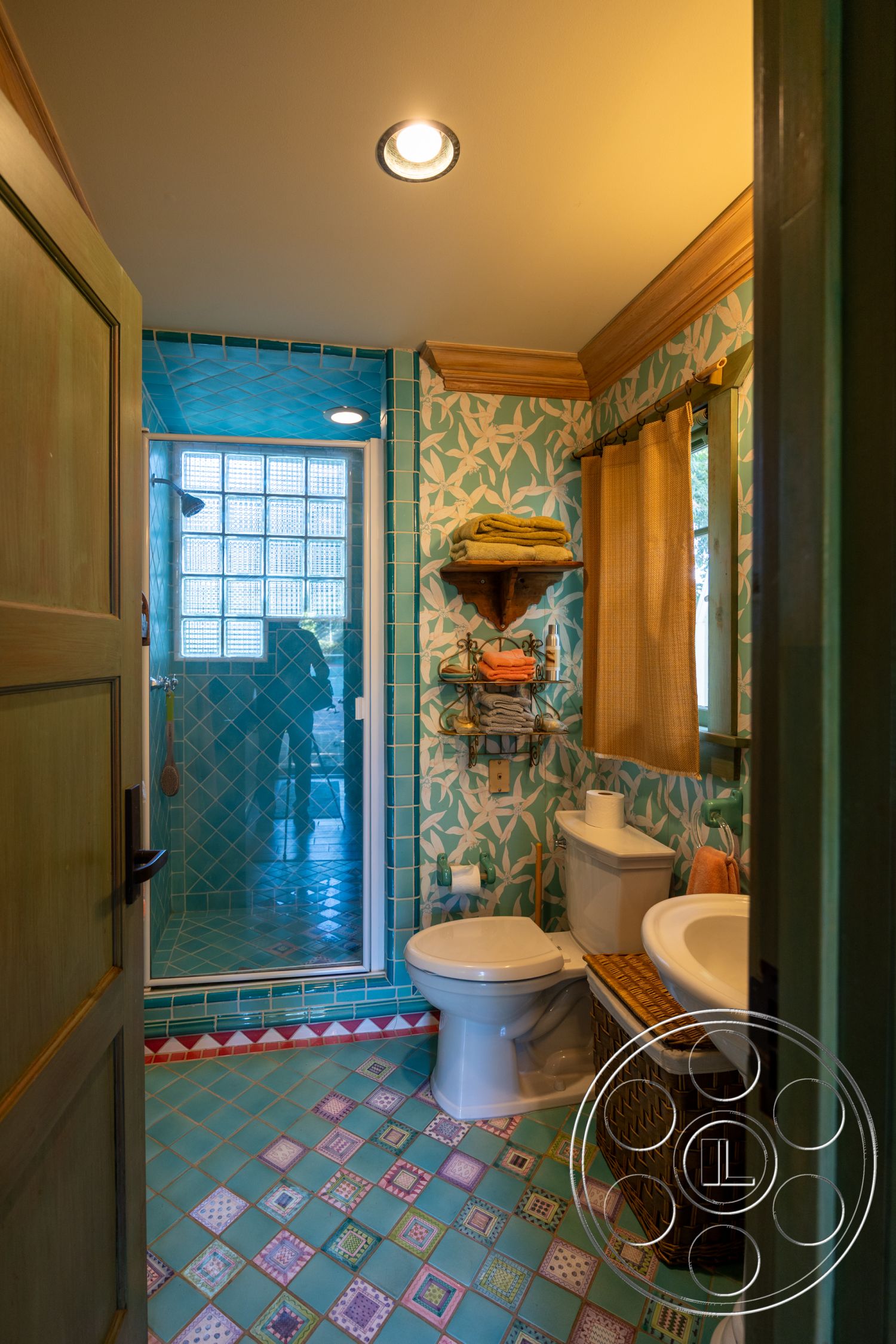 Americana 48 - floral wallpaper, white porcelain toilet, woven storage basket, glass shower doors, modern recessed lighting, tile shower walls, patterned tile bathroom flooring, blue mosaic wall tiles, wooden shelf for towels, decorative fabric shower curtain, green painted door