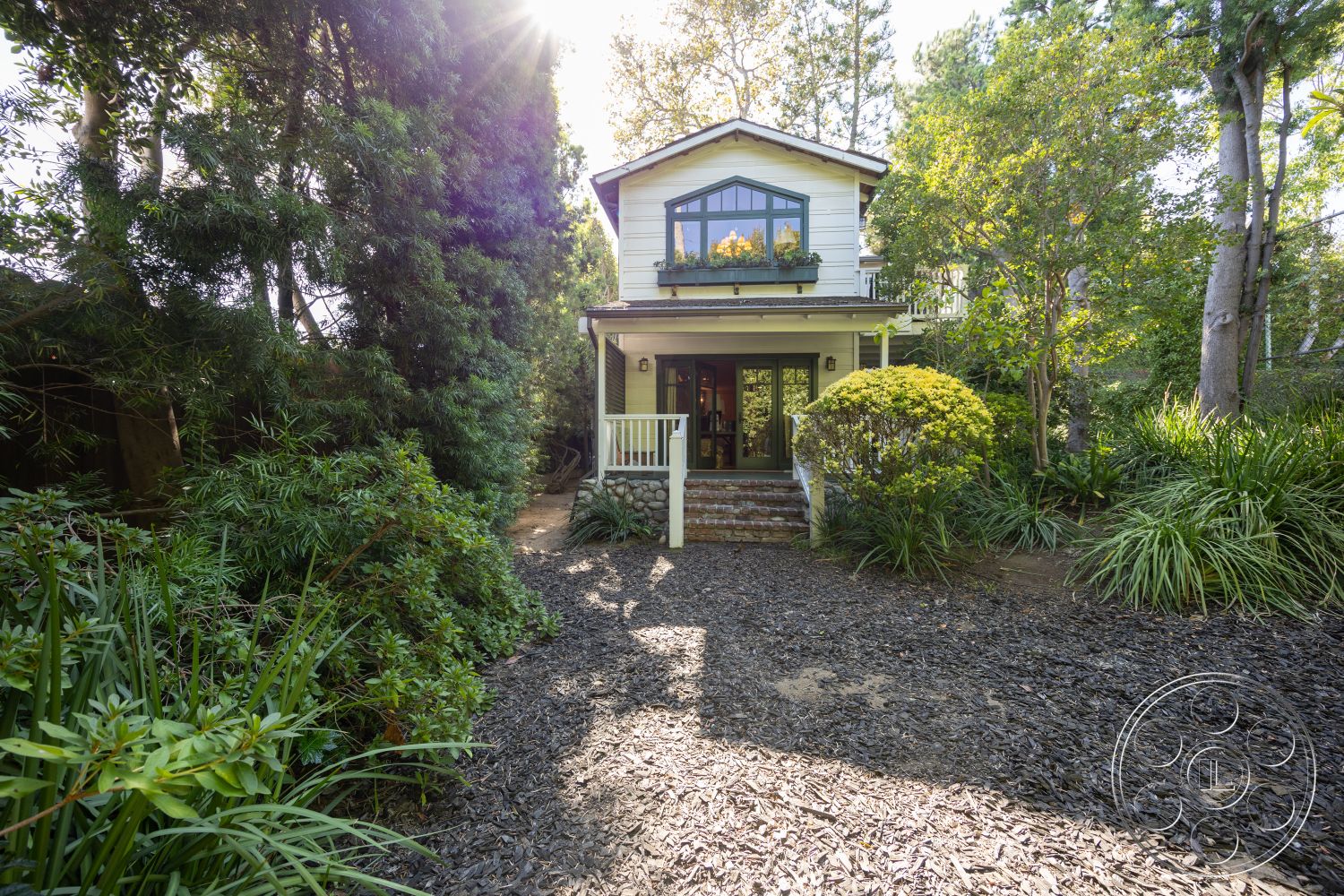 Americana 48 - natural light, exterior, landscaped garden, lush greenery, gabled roof, tall trees, wood siding, wooden front door, rectangular windows, two-story home, front porch with railing, stone front steps, built-in flower boxes, mulch pathway, bright exterior with white paint