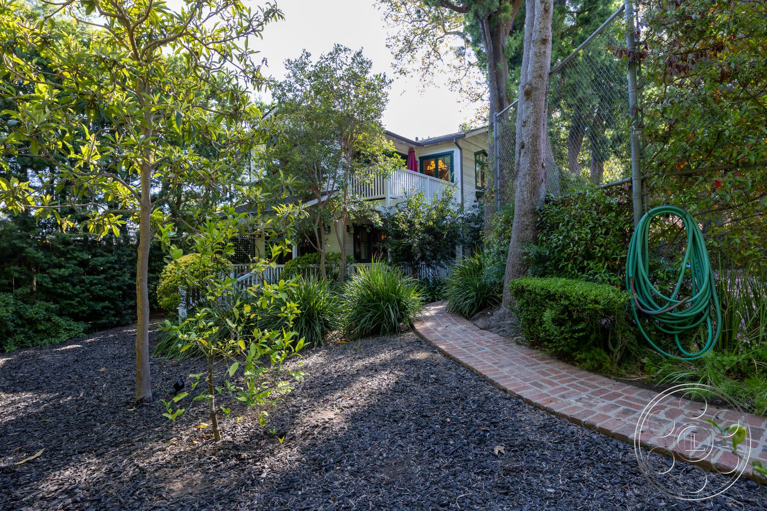 Americana 48 - exterior, brick pathway, residential backyard, garden landscaping, deciduous trees, mulch ground cover, two-story home, outdoor garden space, lush green shrubs, wooden balcony railing