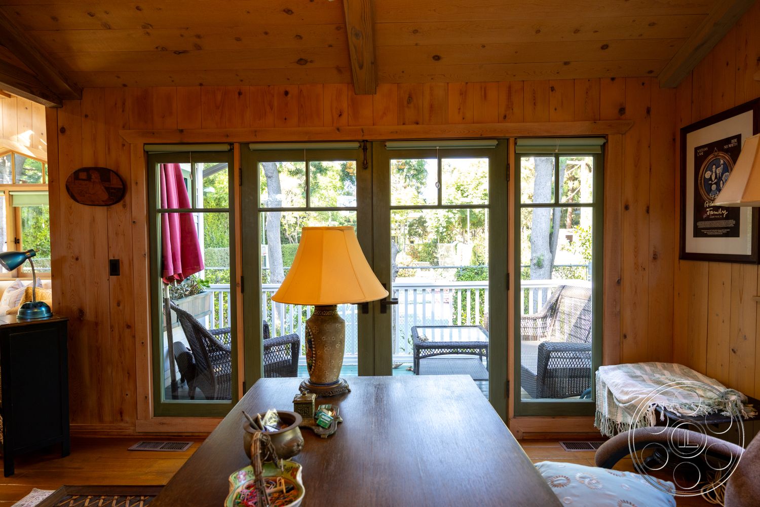 Americana 48 - hardwood flooring, artwork on walls, textured throw blanket, decorative table lamp, exposed wood ceiling beams, wood paneled walls, rustic wooden table, large windows with green frames, woven armchairs, green accent curtains