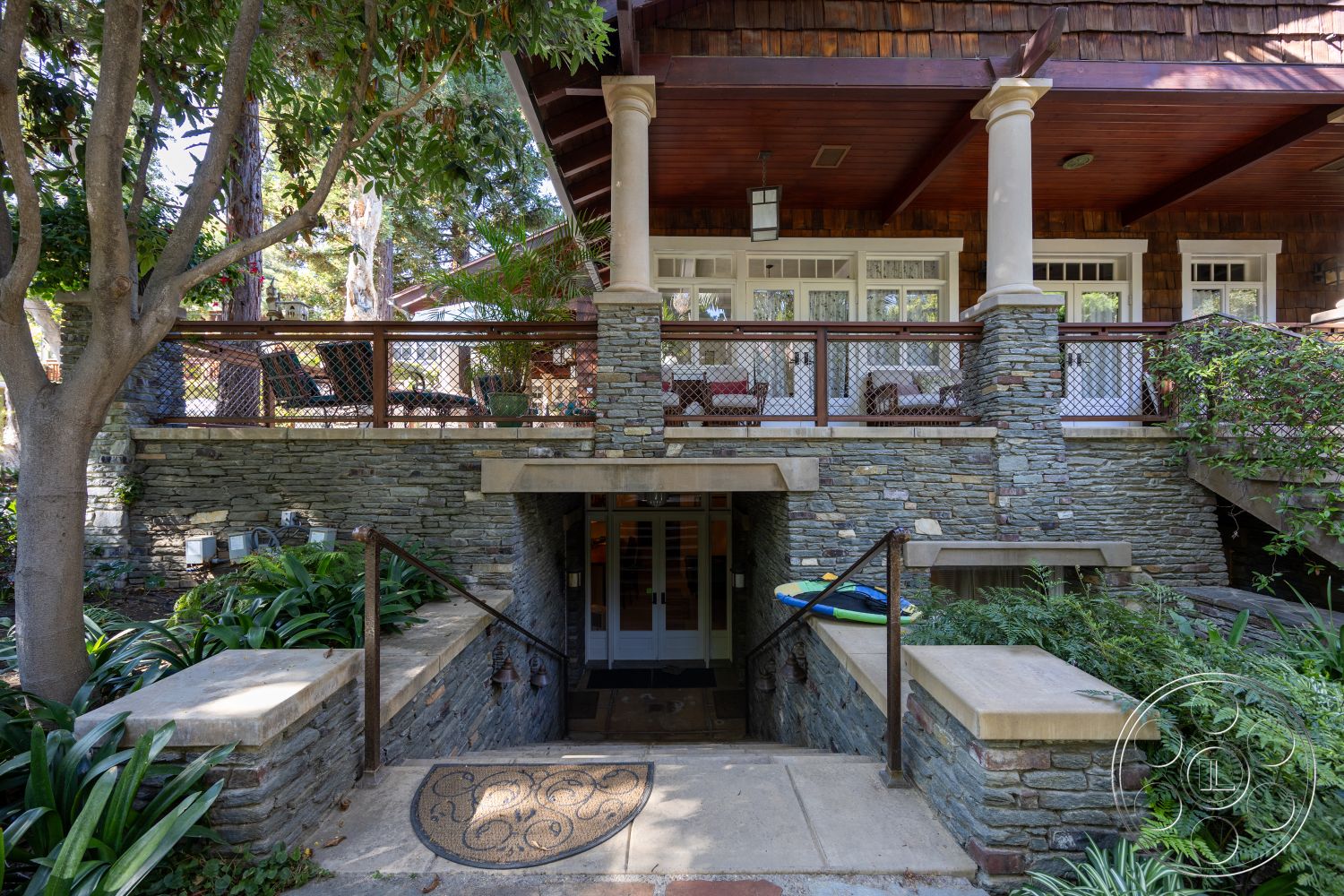 Americana 48 - exterior, landscaped garden, outdoor seating area, surrounding trees, paved walkway, wooden railing, traditional architectural style, stone steps, stone retaining wall, double doors entrance, wooden porch ceiling, exposed timber beams