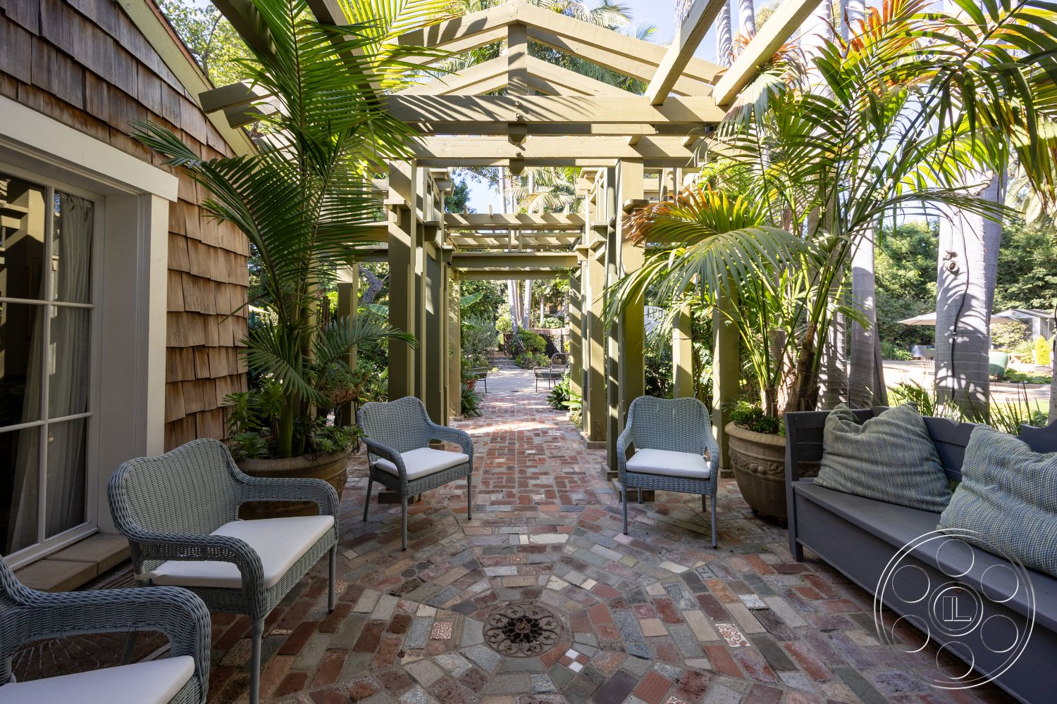 Americana 48 - exterior, outdoor seating area, green foliage, shingle siding, tropical landscaping, planter pots, potted palm trees, brick pathway flooring, open trellis structure, wooden supporting beams, wicker patio chairs, soft outdoor cushions