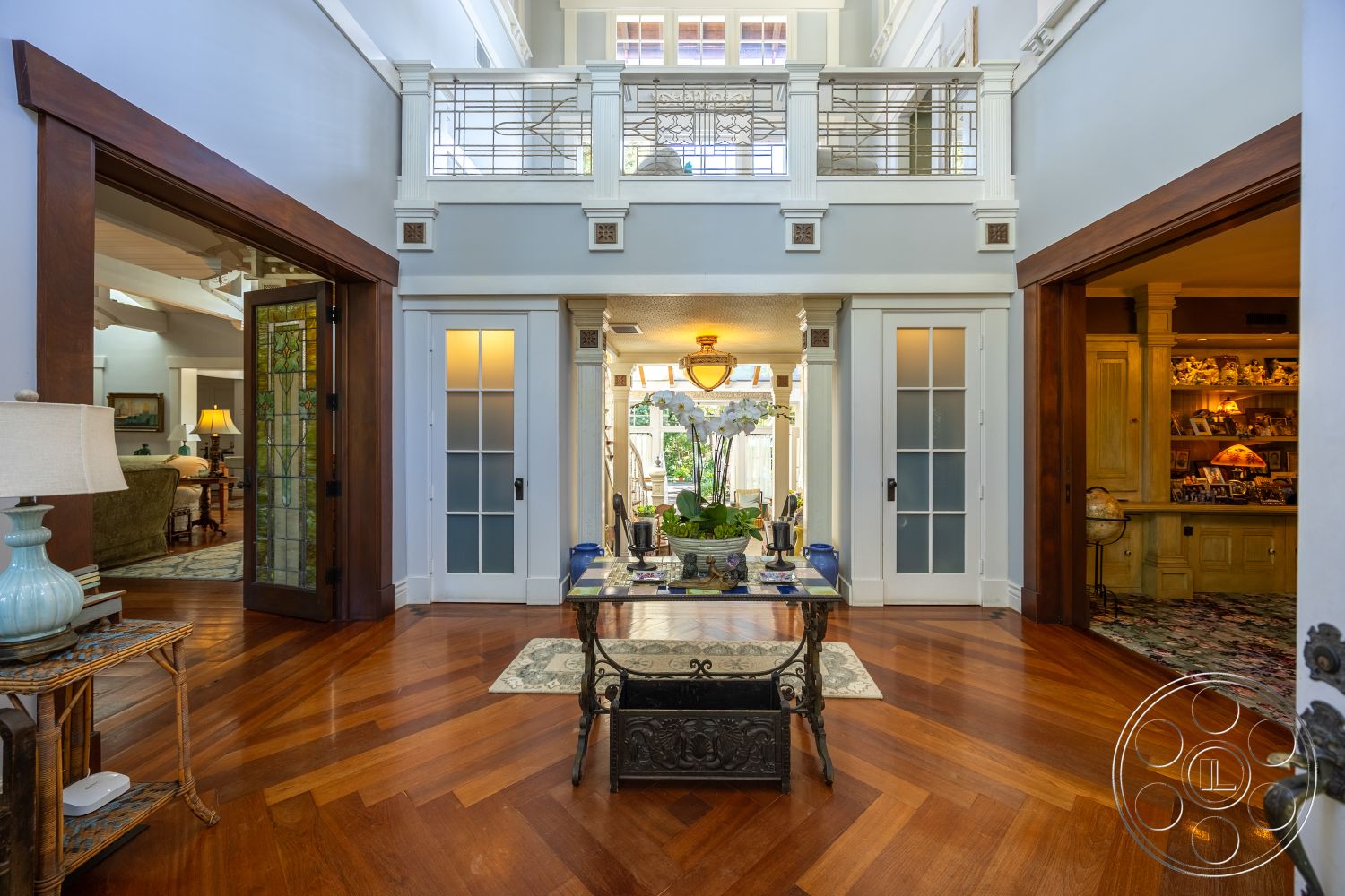 Americana 48 - plant decor, open concept layout, arched doorways, living room access, neutral painted walls, coffered ceiling details, glass panel doors, traditional chandelier lighting, hardwood herringbone flooring, built-in wooden cabinetry, decorative wall moldings, stained glass door panel, spiral staircase railings, tall vaulted ceiling, ornate light fixture, foyer area, framed art displays