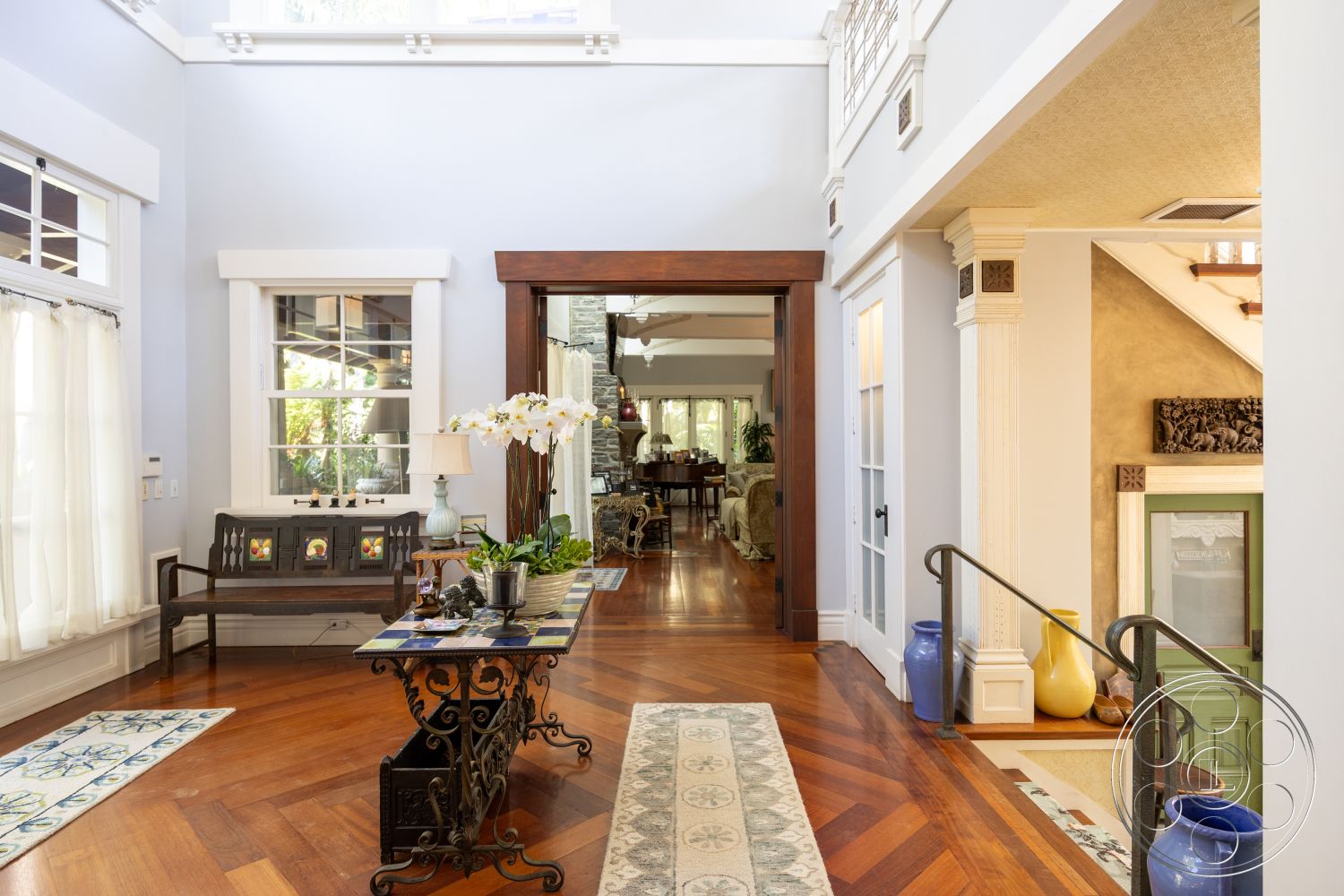 Americana 48 - stone accent wall, exposed beam ceiling, decorative wall art, decorative crown molding, white painted walls, large windows providing natural light, vaulted ceiling with skylights, ornamental column details, hardwood entryway floor, intricate wood staircase, traditional wooden door frame, brass finish door hardware, dining area view through doorway, detailed wood paneling, potted plants and floral arrangements, textile area rugs with geometric patterns, modern black metal console table