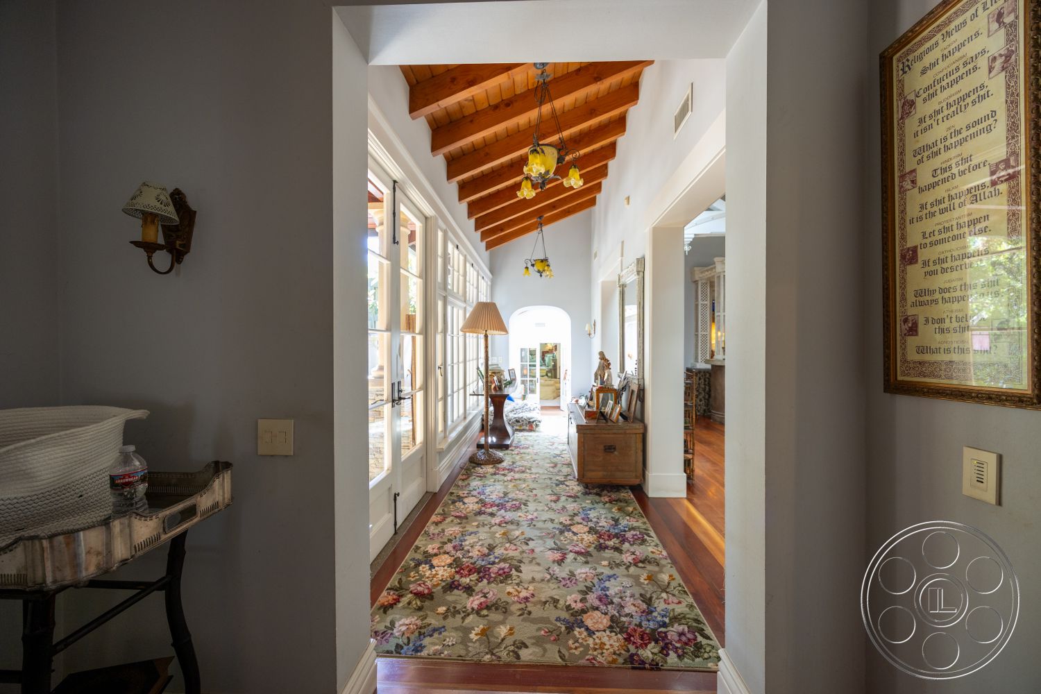 Americana 48 - decorative wall art, arched doorways, traditional wall sconces, exposed wood ceiling beams, interior hallway, wooden furniture accents, natural light filled space, textured wall paint, hardwood floor hallway, floral area rug, windowed entryway