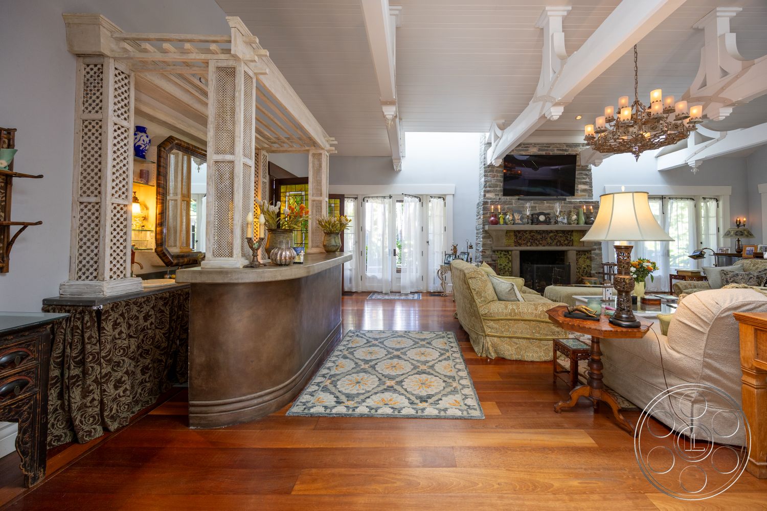 Americana 48 - natural light, open floor plan, glass cabinet doors, textured plaster walls, decorative table lamp, chandelier light fixture, exposed wood ceiling beams, hardwood living room floors, stone fireplace wall, white painted ceiling, curved bar countertop, floral area rug, traditional style furniture, classic home decor