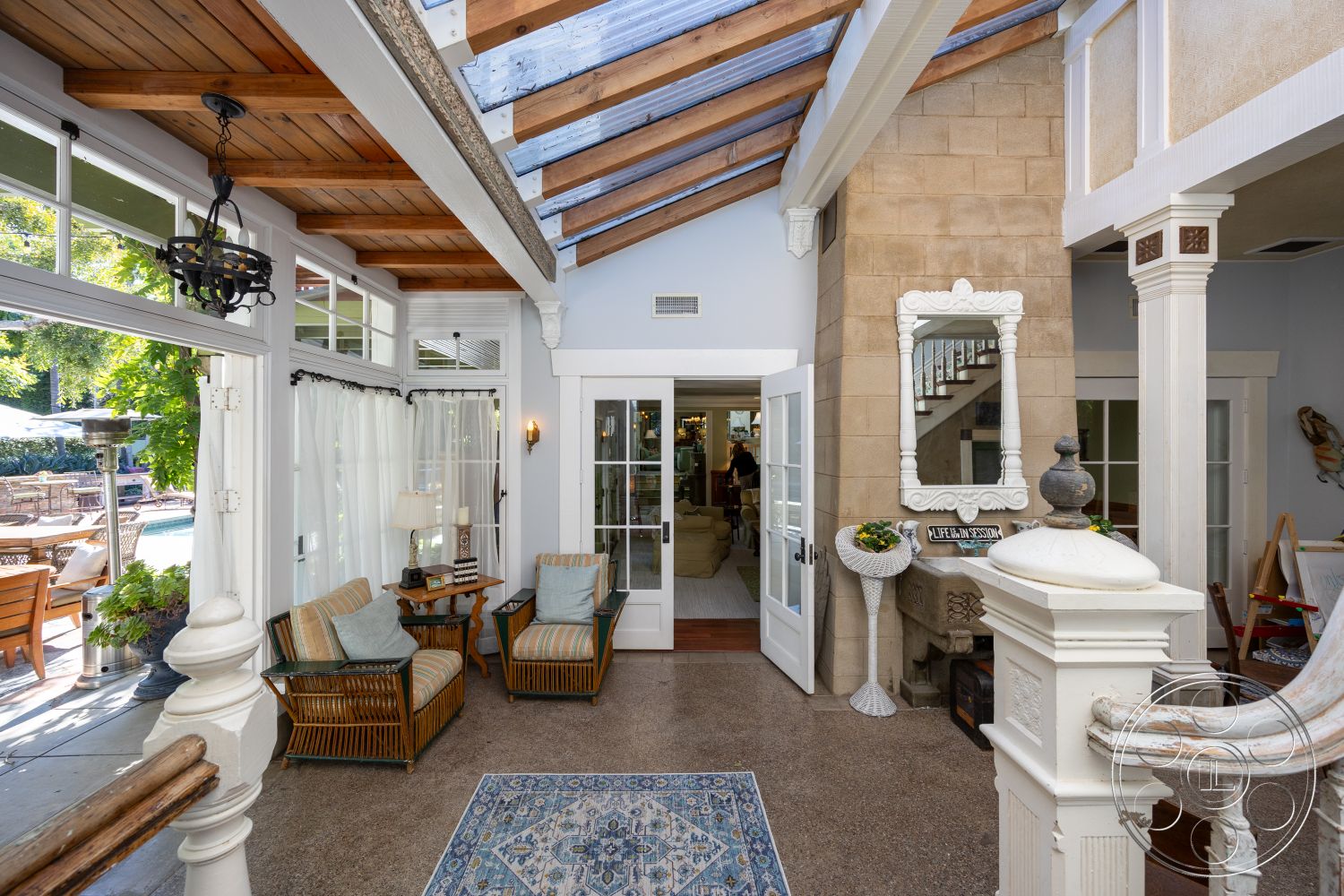 Americana 48 - natural light, stone accent wall, potted plants, transom windows, glass sliding doors, open concept design, striped area rug, hardwood floor, outdoor view, textured plaster walls, wooden accent furniture, wrought iron chandelier, exposed wood ceiling beams, decorative wall mirror, curtained window treatments, white framed windows, casual sitting area