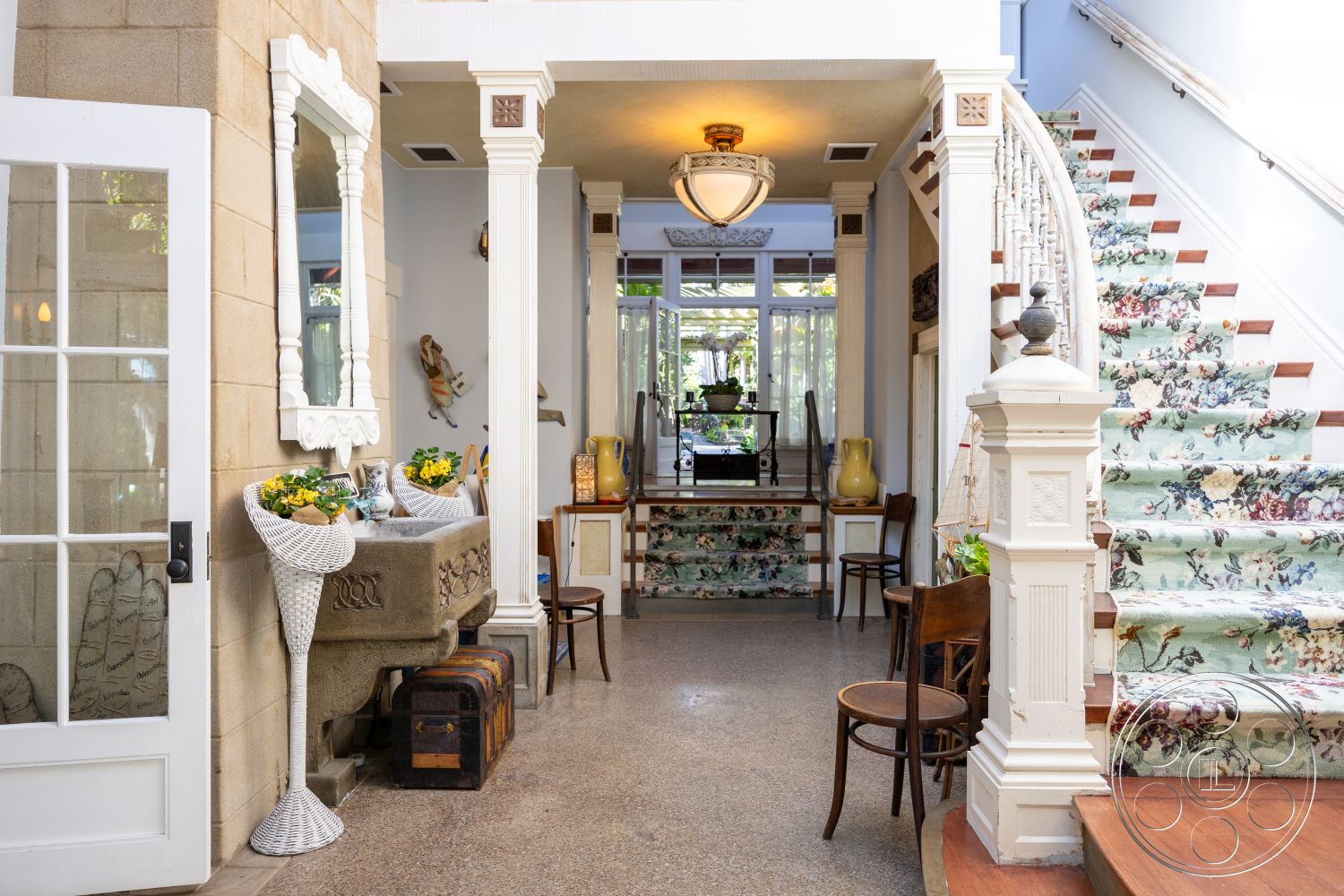 Americana 48 - textured plaster walls, decorative ceramic vase, open view to outdoor space, coffered ceiling details, traditional chandelier lighting, decorative wall mirror, stone accent walls, built-in wooden cabinetry, curved wooden staircase, granite entrance floor, floral patterned carpet stairs, white wooden balustrade, vintage wooden chair, green potted plants