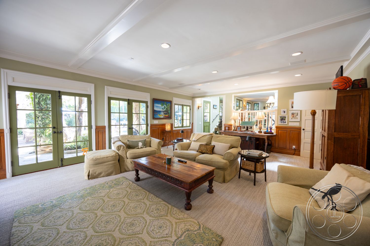 Americana 48 - textured area rug, bright natural light, framed wall art, hardwood floor, warm wood paneling, home office nook, exposed wood ceiling beams, white crown molding, decorative table accessories, green painted walls, traditional wooden coffee table, upholstered beige sofa, green double French doors, wooden built-in cabinetry, light fixtures with lampshades, seating area with accent pillows, open layout living room, family room setting