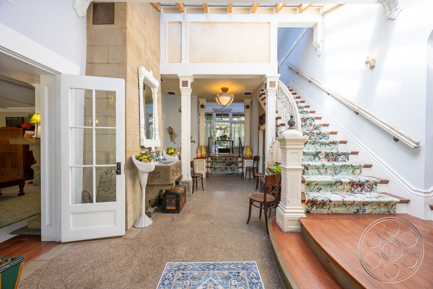 Americana 48 - exterior, decorative ceiling beams, bright natural light from windows, elegant chandelier lighting, wood trim accents, terrazzo floor entryway, curved staircase with floral carpet, wooden steps with white risers, cream-colored stone wall, arched doorways for entry, white wooden double doors, framed mirror with ornate design, floral accent area rug, accessible leading to main living area