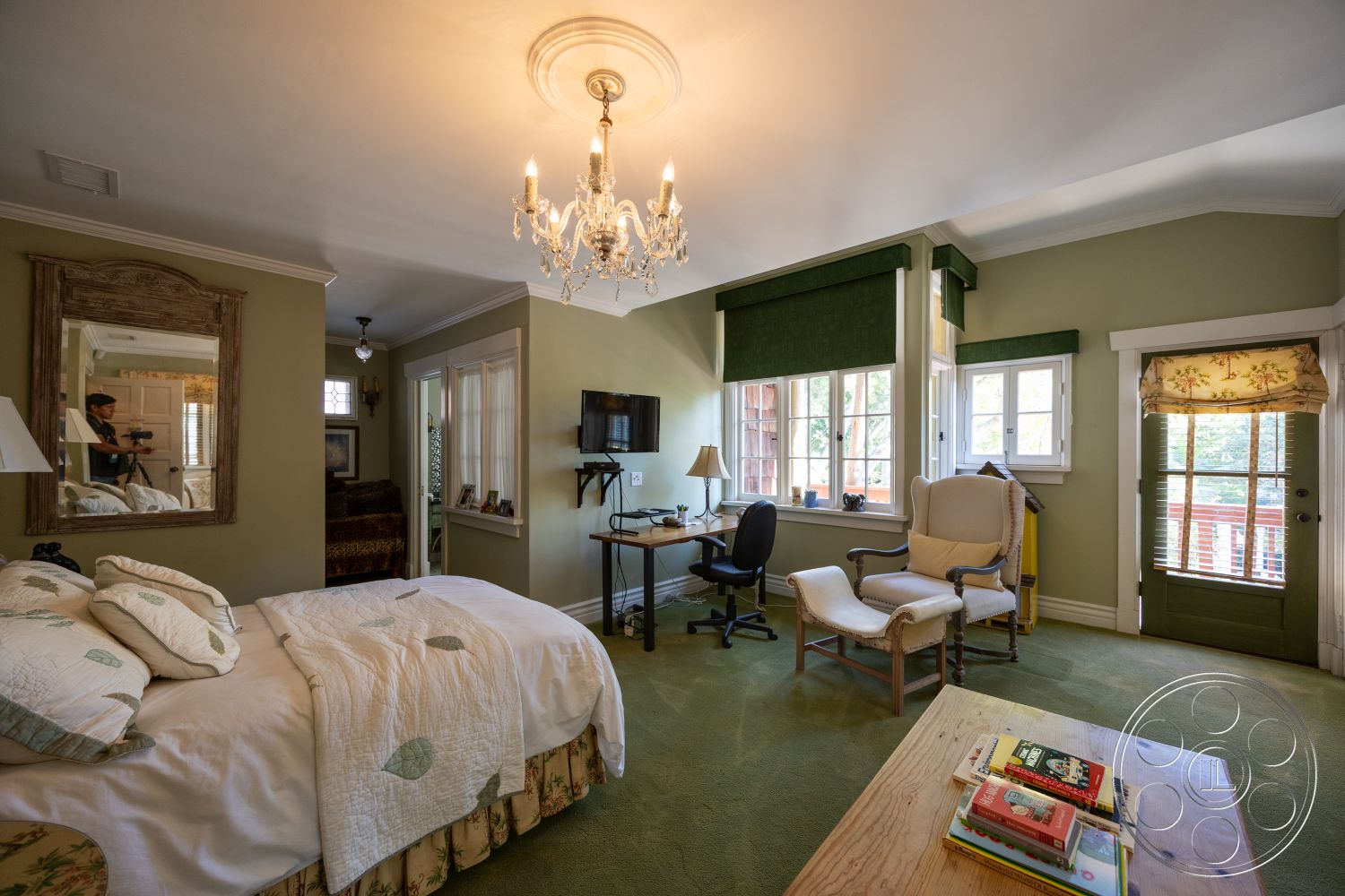 Americana 48 - wall-mounted television, wooden bedside table, arched doorways, upholstered armchair, mirror with decorative frame, natural light from multiple windows, decorative accent pillows, carpet bedroom flooring, crystal chandelier lighting, green painted walls, wooden ceiling trim, bi-fold window treatments, traditional style bedroom, desk area with office chair