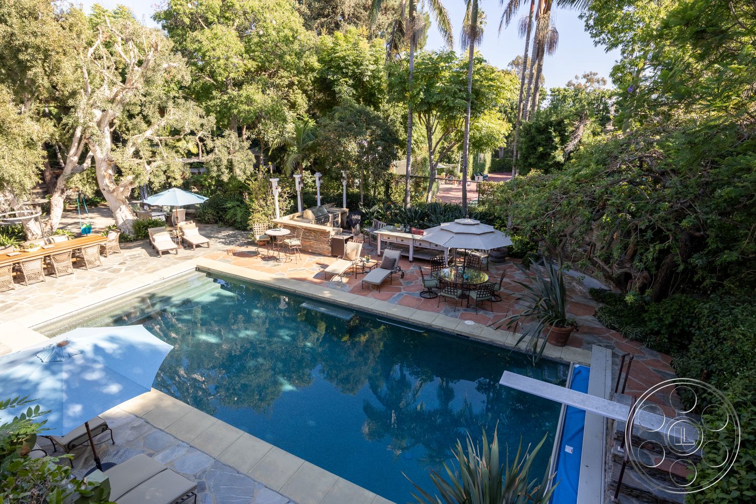 Americana 48 - exterior, natural landscaping, stone barbecue area, paved walkway, metal patio chairs, surrounding palm trees, stone pool deck, rectangle swimming pool, lush garden setting, concrete pool border, terracotta patio flooring, wooden outdoor dining table, canvas umbrella