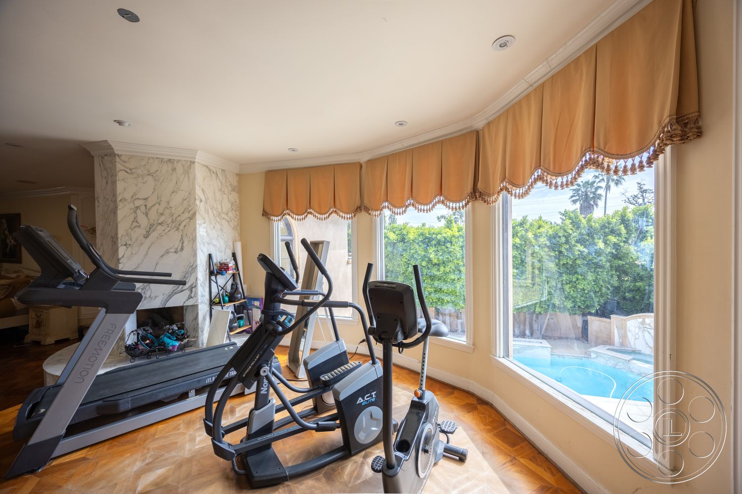 Mansion 146 - hardwood flooring, treadmill, pool view, elliptical machine, marble feature wall, decorative window treatment, crown molding details, large bay windows, luxury fitness room, lush backyard landscaping