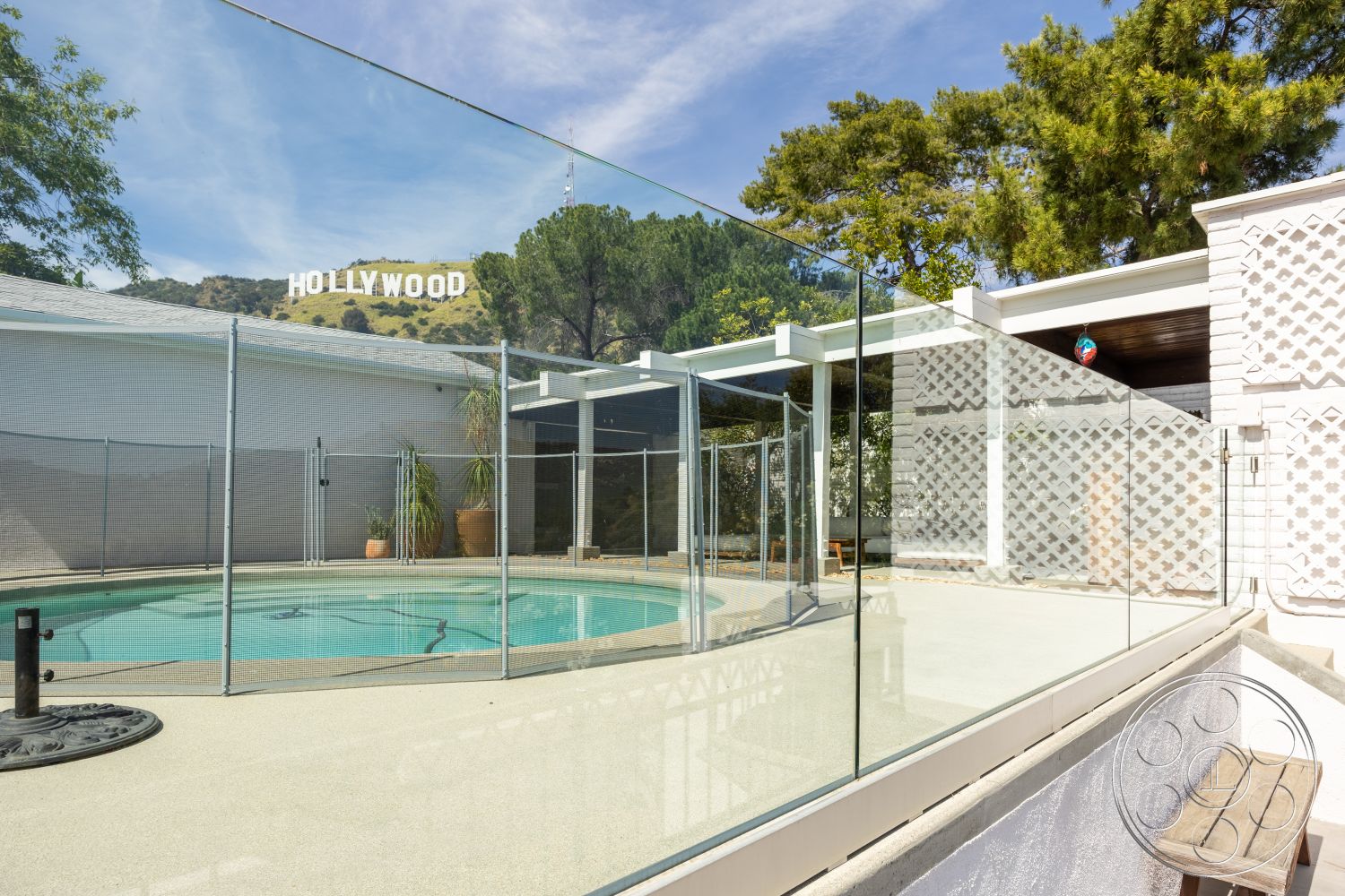 Retro 288 - exterior, oval pool, lush greenery surroundings, concrete patio flooring, textured plaster wall, concrete pool border, horizontal wood slat details, glass pool safety fence, Hollywood sign view, stone potted plant features, bright sunny atmosphere