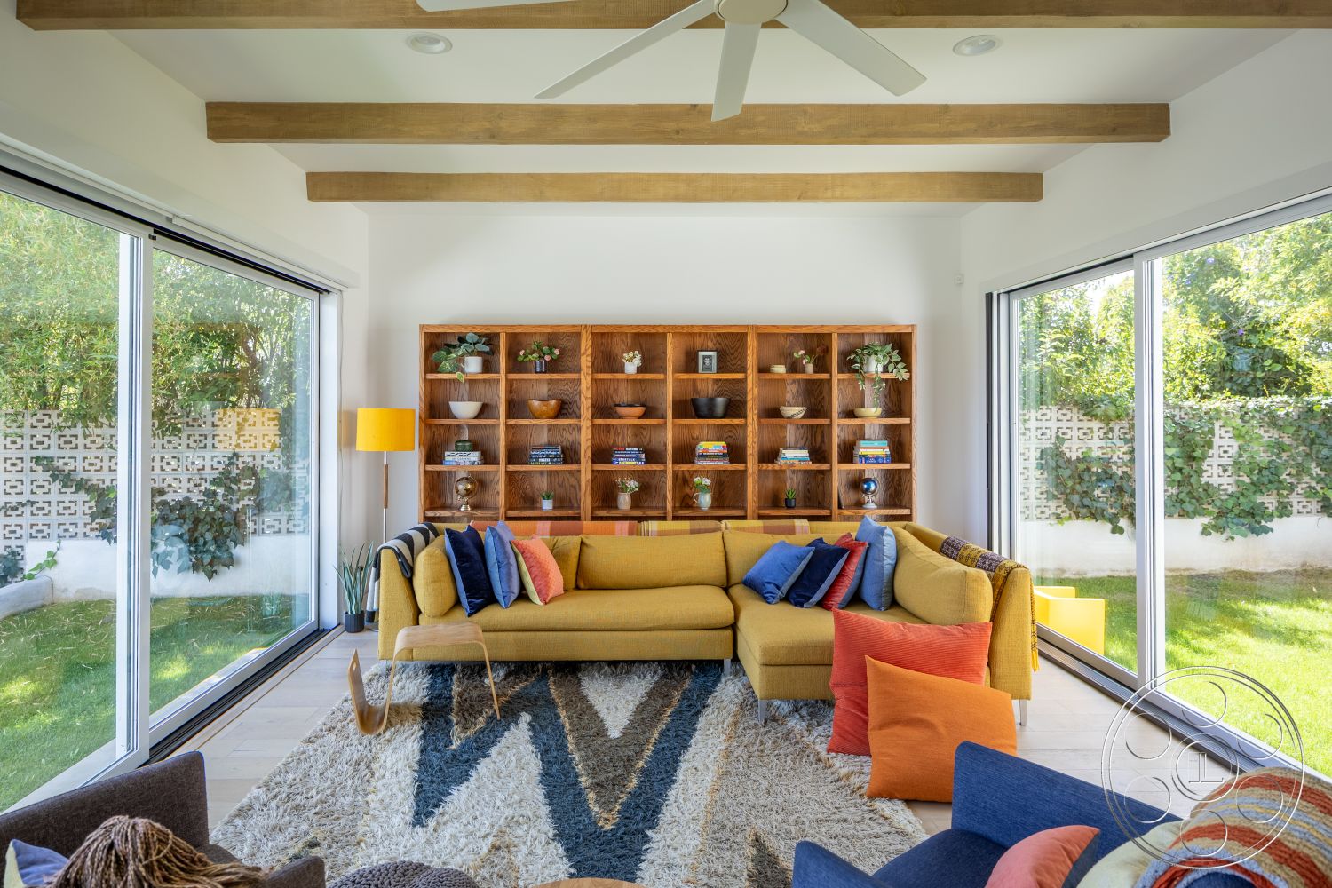 Retro 288 - light wood flooring, large glass windows, colorful throw pillows, comfortable seating area, greenery view through windows, contemporary lighting fixture, exposed wood ceiling beams, white painted walls, cozy sectional sofa, modern area rug, built-in wooden shelving unit