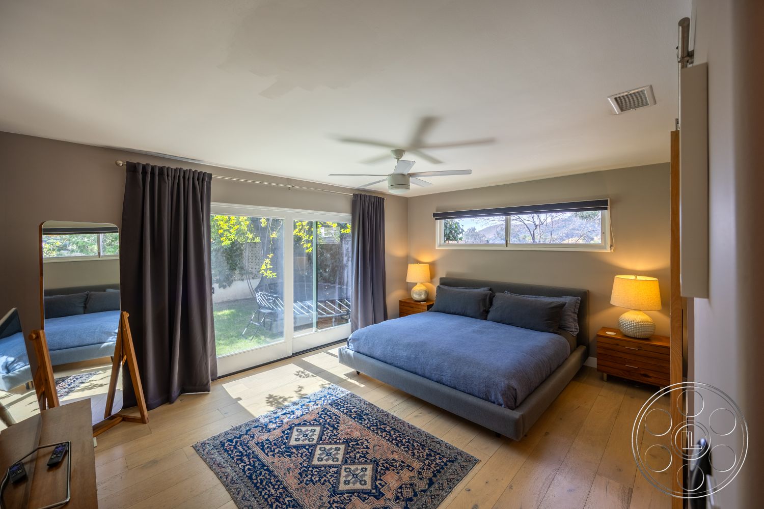 Retro 288 - natural light from windows, wooden bedside tables, light-colored curtains, wall-mounted mirror, hardwood bedroom floors, floor-to-ceiling sliding glass doors, contemporary minimalist design, gray painted walls, flat ceiling with ceiling fan, modern decorative area rug, blue upholstered bed, matching table lamps