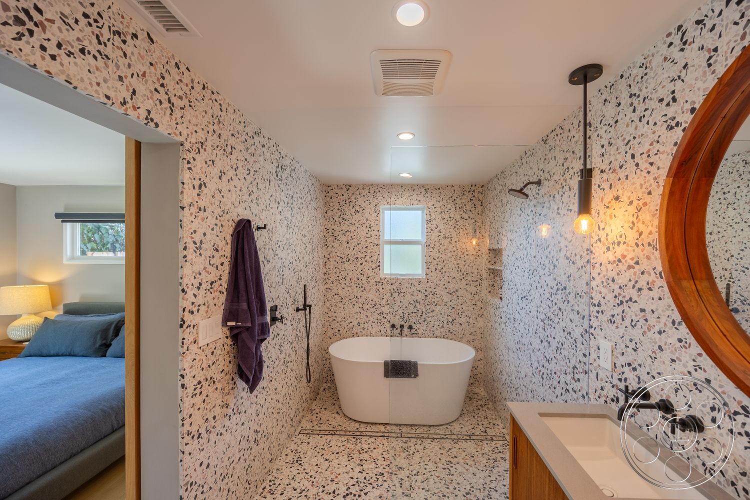 Retro 288 - neutral color palette, textured wall treatment, natural light through window, minimalist bathroom design, contemporary bathroom aesthetic, freestanding soaking tub, terrazzo tile bathroom walls, black wall-mounted faucet, wooden round mirror, transparent glass shower partition, light fixture pendant lighting, wood cabinetry with sleek lines, decorative towel hanging rack