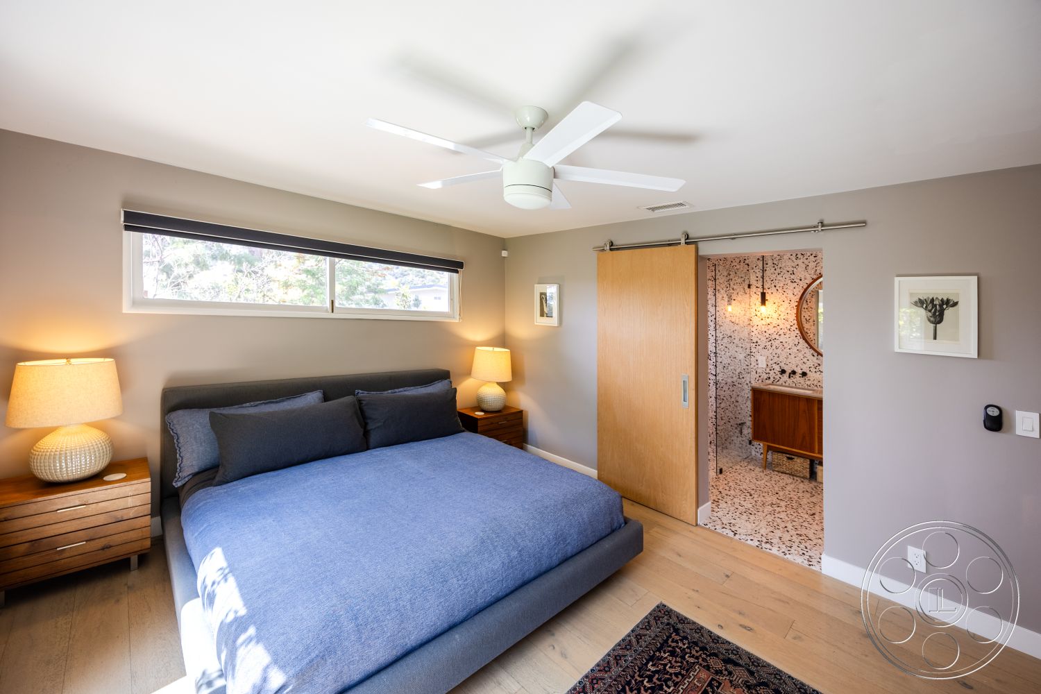 Retro 288 - round mirror, floor-to-ceiling windows, soft ambient lighting, modern ceiling fan, hardwood bedroom floors, terrazzo bathroom flooring, gray painted walls, walnut nightstands, textured blue bedding, sliding wooden barn door, minimalist wood vanity