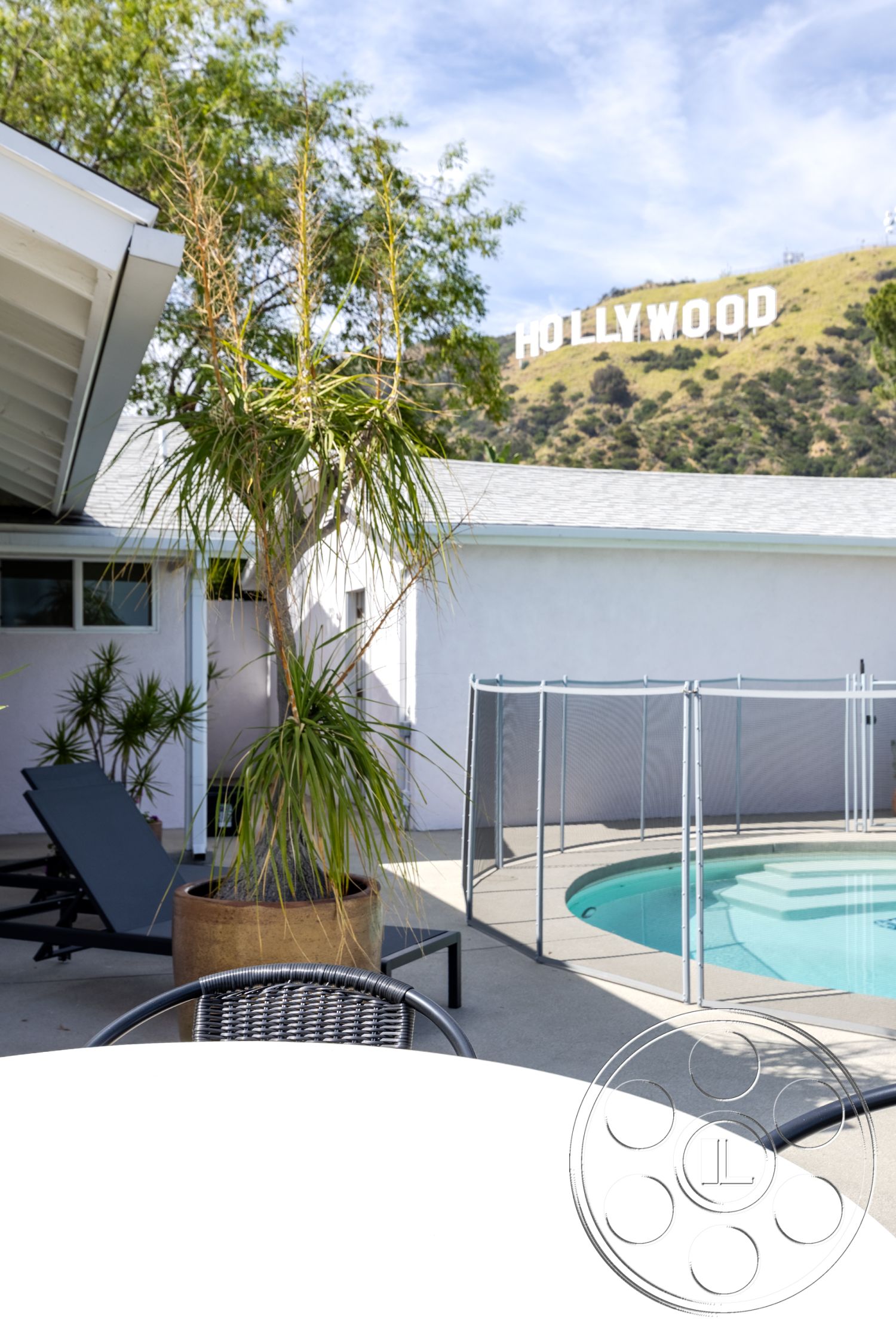 Retro 288 - exterior, glass pool fence, rectangular pool, concrete patio floor, planter with tropical plants, Hollywood sign view, supporting walls with smooth white finish, outdoor lounge chair, circular outdoor table, desert landscape backdrop