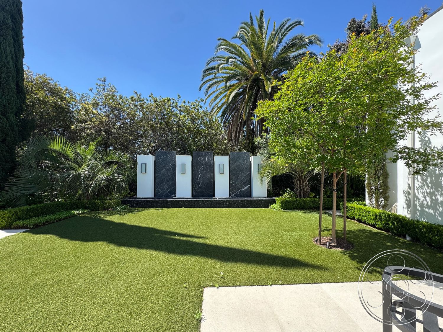 Modern 522 - exterior, white stucco walls, lush greenery, palm trees, modern water feature, tropical landscaping, artificial turf lawn, concrete patio floor, dark stone feature wall, rectangular outdoor layout
