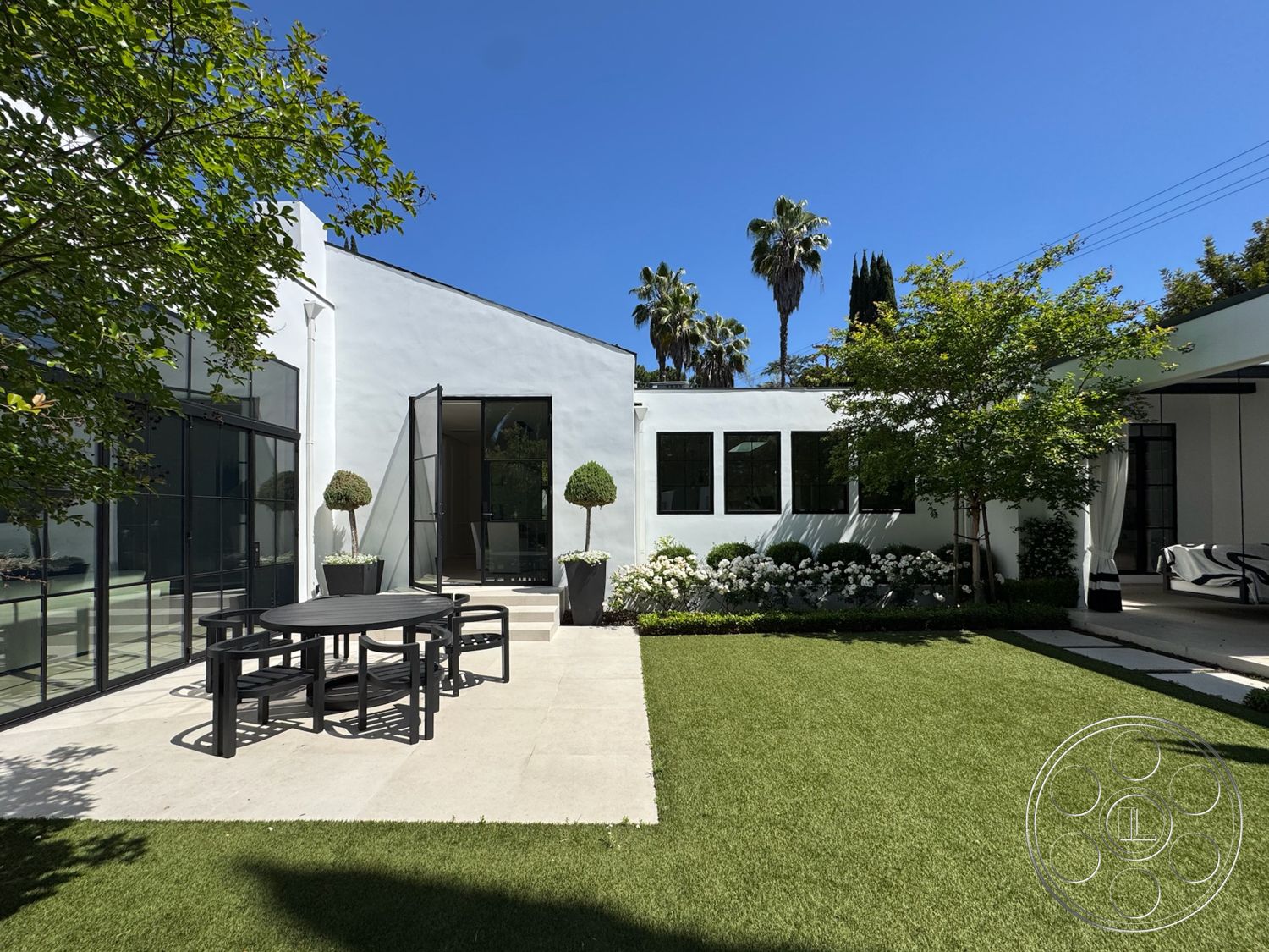Modern 522 - exterior, landscaped garden, white stucco walls, modern architecture, palm trees, lush green grass, rectangular dining table, concrete patio flooring, metal chair design, symmetrical landscape design, large black-framed windows, hardscaped outdoor area, architectural glass doors, drought-resistant planting