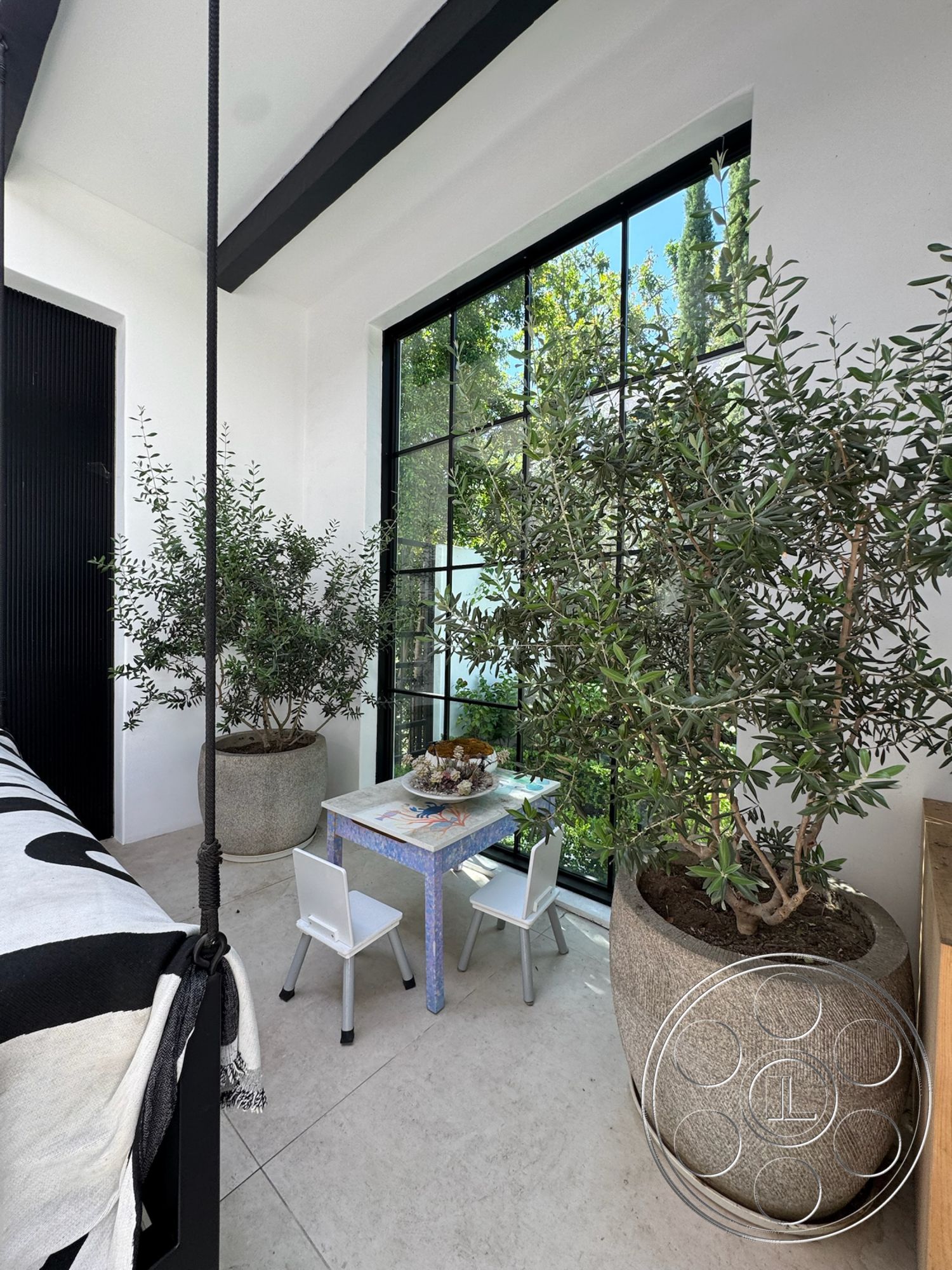 Modern 522 - polished concrete flooring, exposed wood ceiling beams, large steel framed windows, light-colored plaster walls, modern children's table, minimalist white chairs, large round stone planters, green potted olive trees, decorative tabletop arrangement