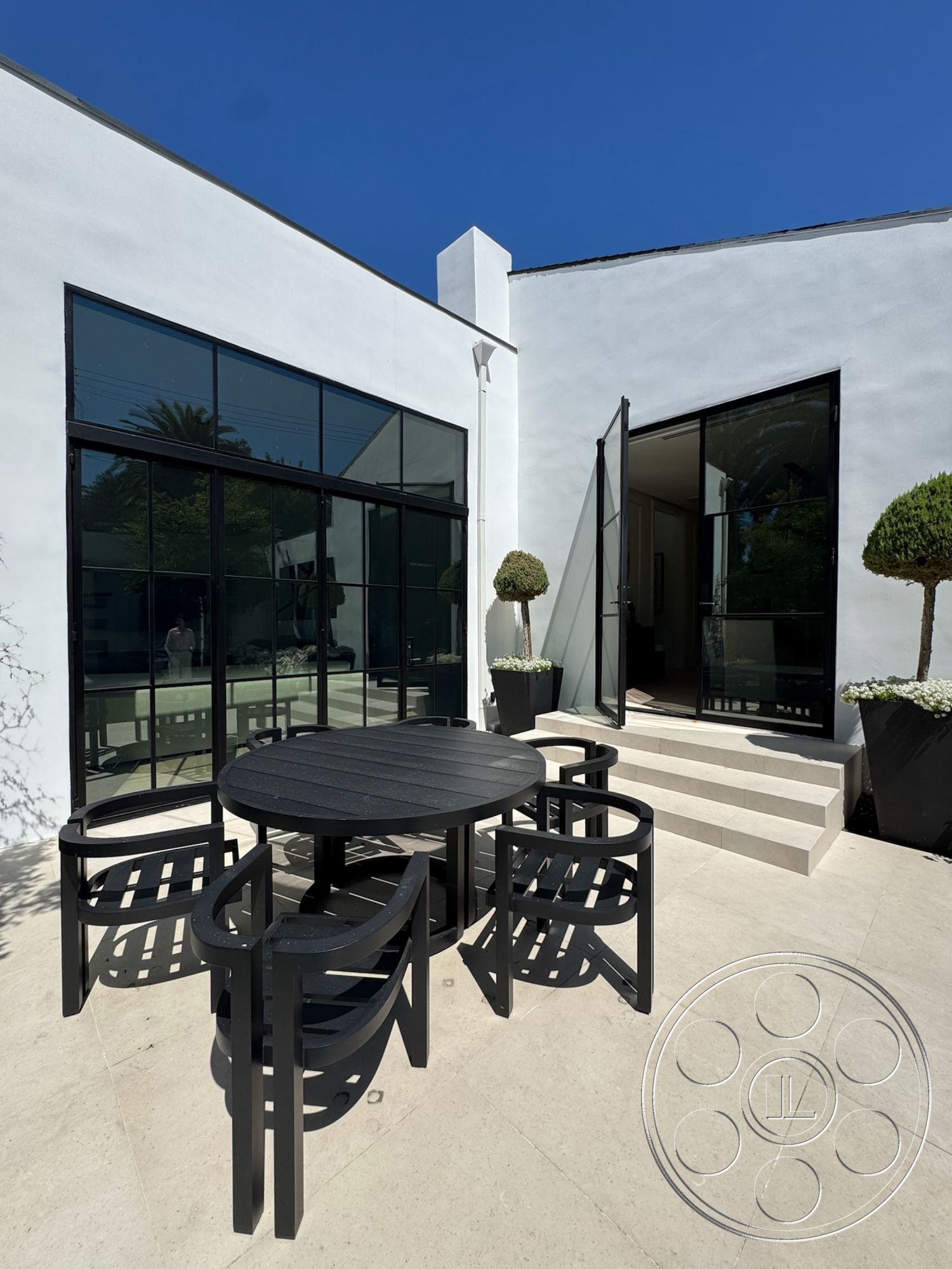 Modern 522 - exterior, white stucco walls, floor-to-ceiling windows, modern architectural style, black outdoor furniture, rectangular dining table, concrete patio flooring, glass door entry, geometric patio layout, planter boxes with flowers, potted topiary trees