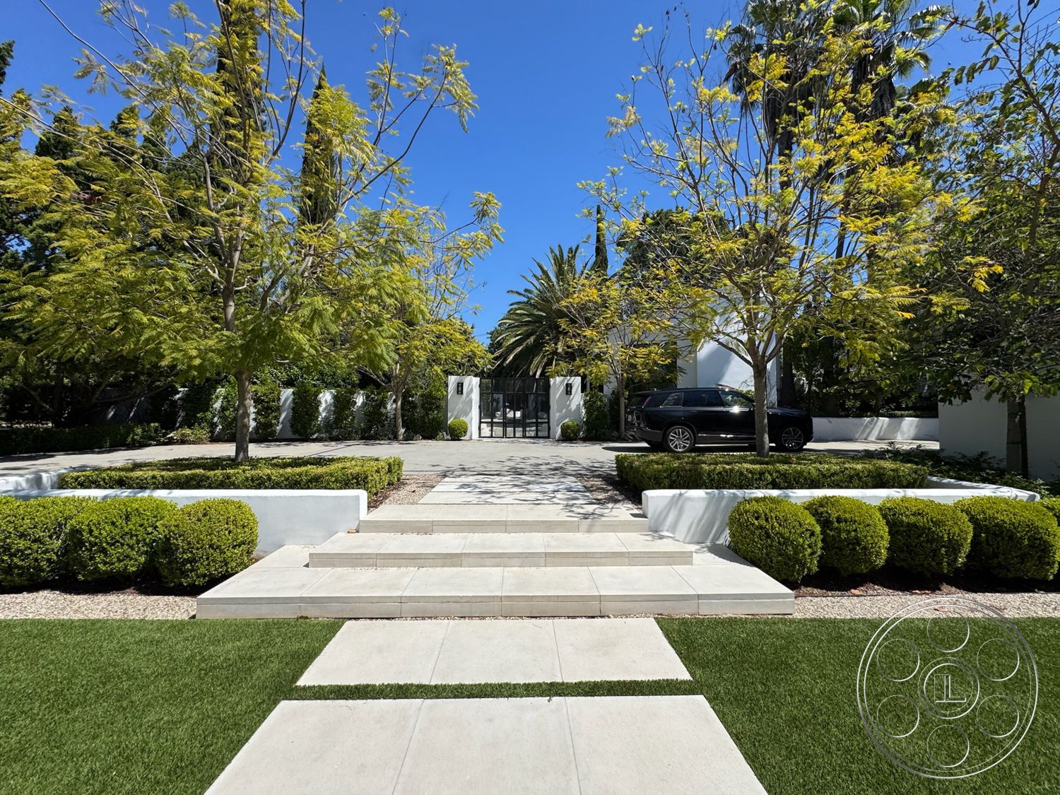 Modern 522 - exterior, paved driveway, modern landscaping, grass lawn, gated entrance, tall palm trees, symmetrical garden design, white concrete walls, minimalist garden, concrete stepping stones, bright blue sky, landscaped shrubs, decorative pebble pathway, bushy hedges