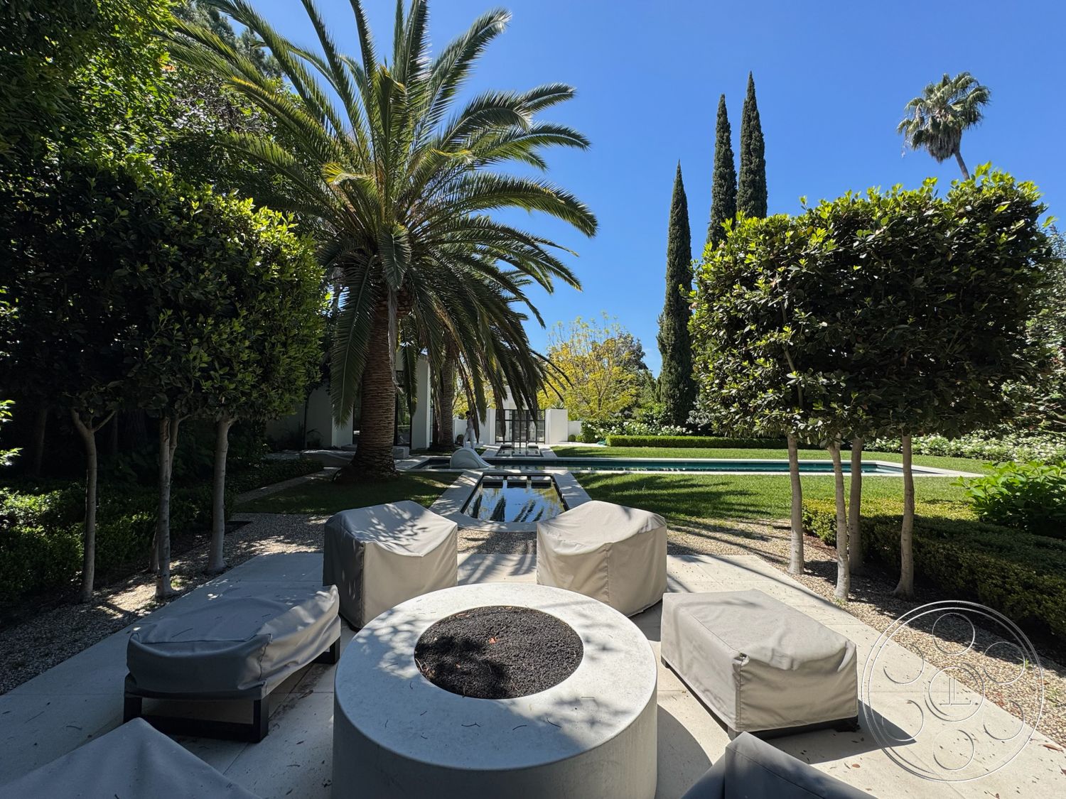 Modern 522 - exterior, palm trees, gravel walkway, tall cypress trees, flat lawn, planted garden beds, concrete patio floor, sunny garden setting, natural stone pool border, modern outdoor lounge chairs, lush green hedge, rectangular stone fire pit