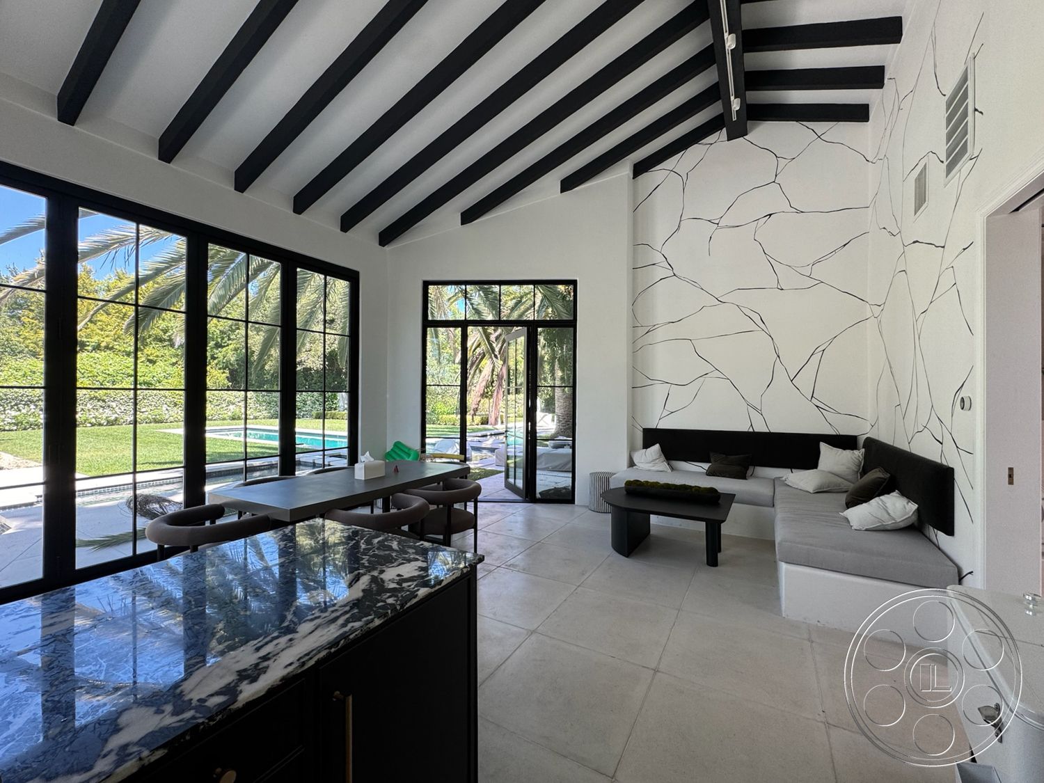 Modern 522 - open layout, black marble countertop, dark wood dining table, natural light flooding the space, vaulted ceiling with black exposed wood beams, large glass panel windows, minimalist concrete flooring, monochrome wall with cracked pattern, L-shaped built-in seating area with gray cushions, indoor-outdoor integration, greenery view from inside, stylish black coffee table