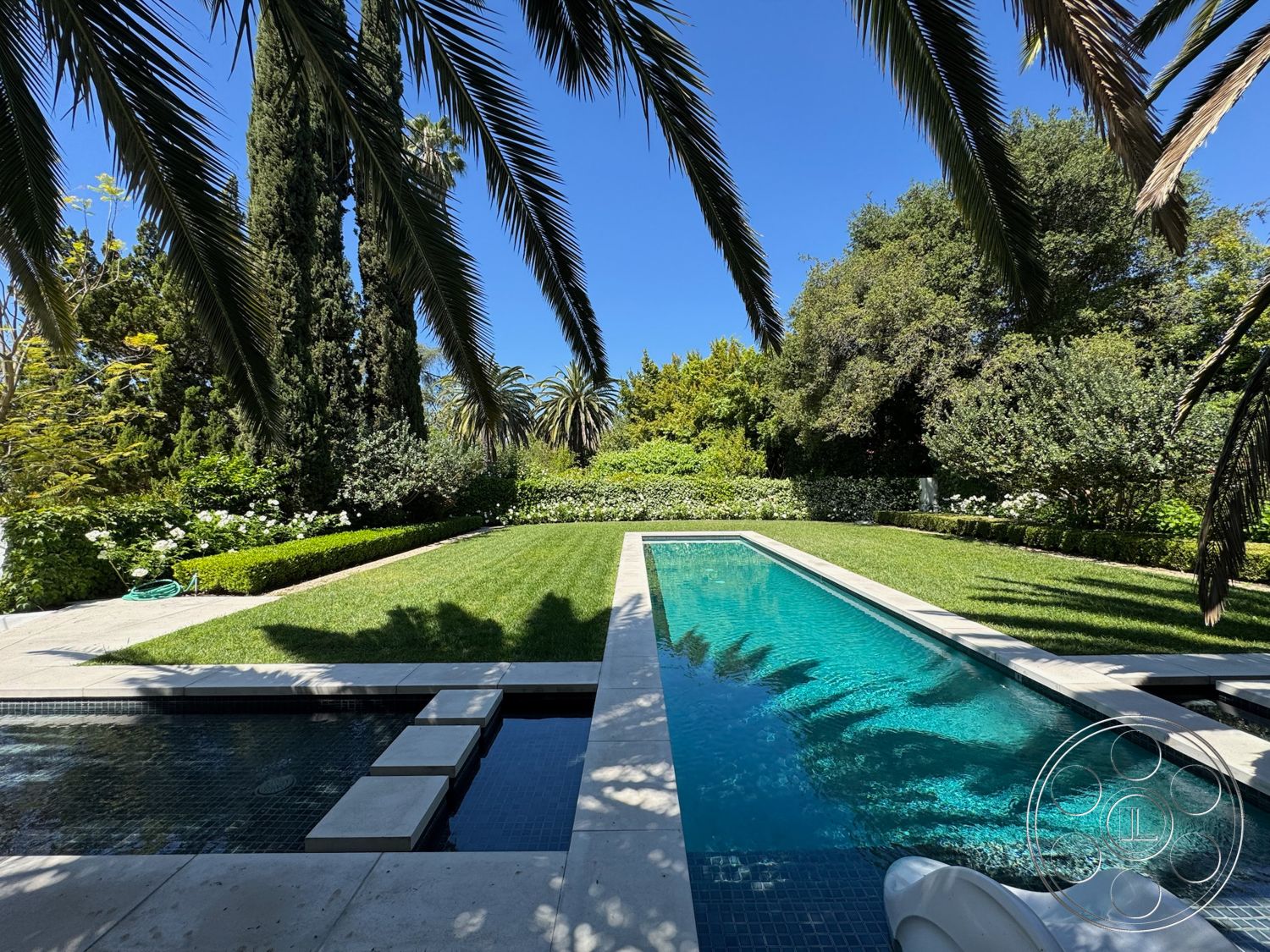 Modern 522 - exterior, water feature, lush green lawn, flowering shrubs, clear blue sky, tropical landscape, tall palm trees, rectangular pool, hedge landscaping, stone patio flooring, concrete pool border, modern minimalist architecture, decorative stone tiles, framed pool decking