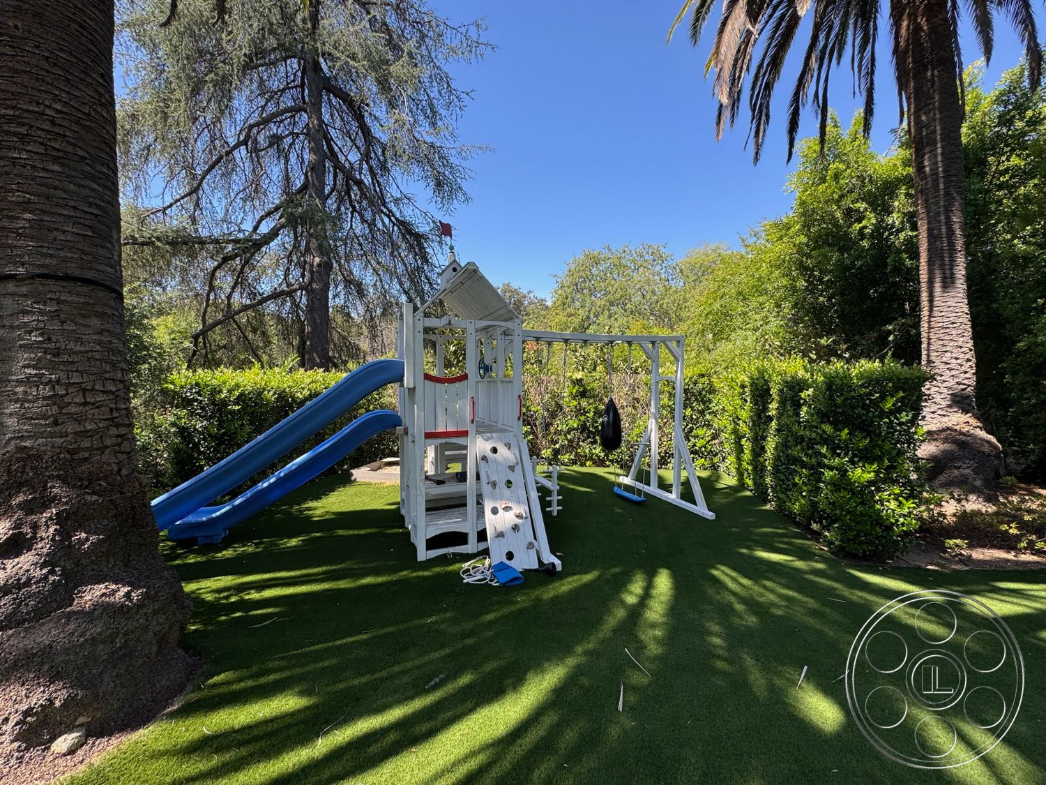 Modern 522 - exterior, tall palm trees, lush green hedges, artificial grass landscaping, sunny backyard, outdoor recreational area, white wooden playset, blue plastic slide, swing set with black seat, rock climbing wall