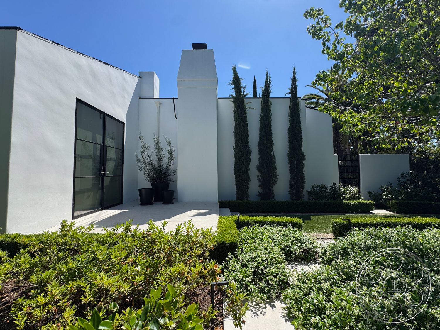 Modern 522 - exterior, white stucco walls, gravel pathway, simple geometric shapes, black metal framed windows, flat roof design, concrete patio flooring, lush green shrubs, outdoor potted plants, minimalistic architectural style, landscape hedges, tall slender cypress trees